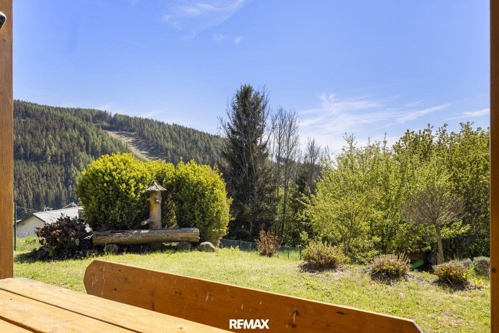 Blick von der Terrasse in den Garten mit grünen Büschen und Bäumen, im Hintergrund Berge.