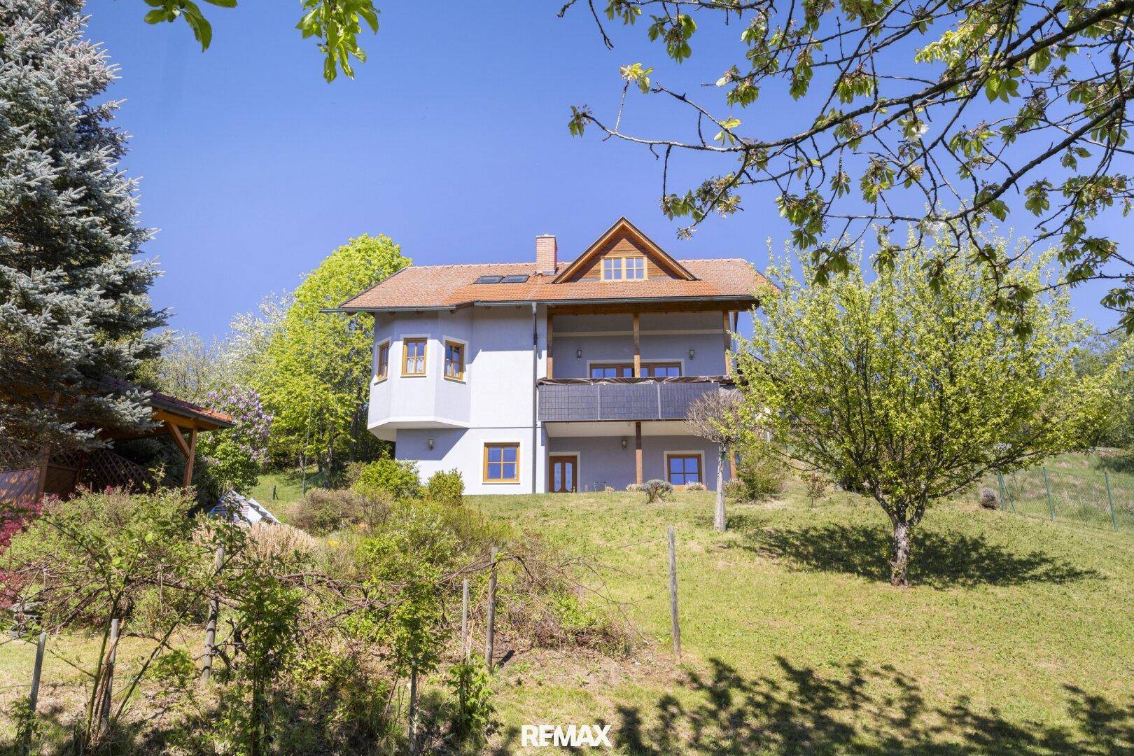Wohnhaus mit heller Fassade, Satteldach und Balkonen, eingebettet in grüne Hügellandschaft.