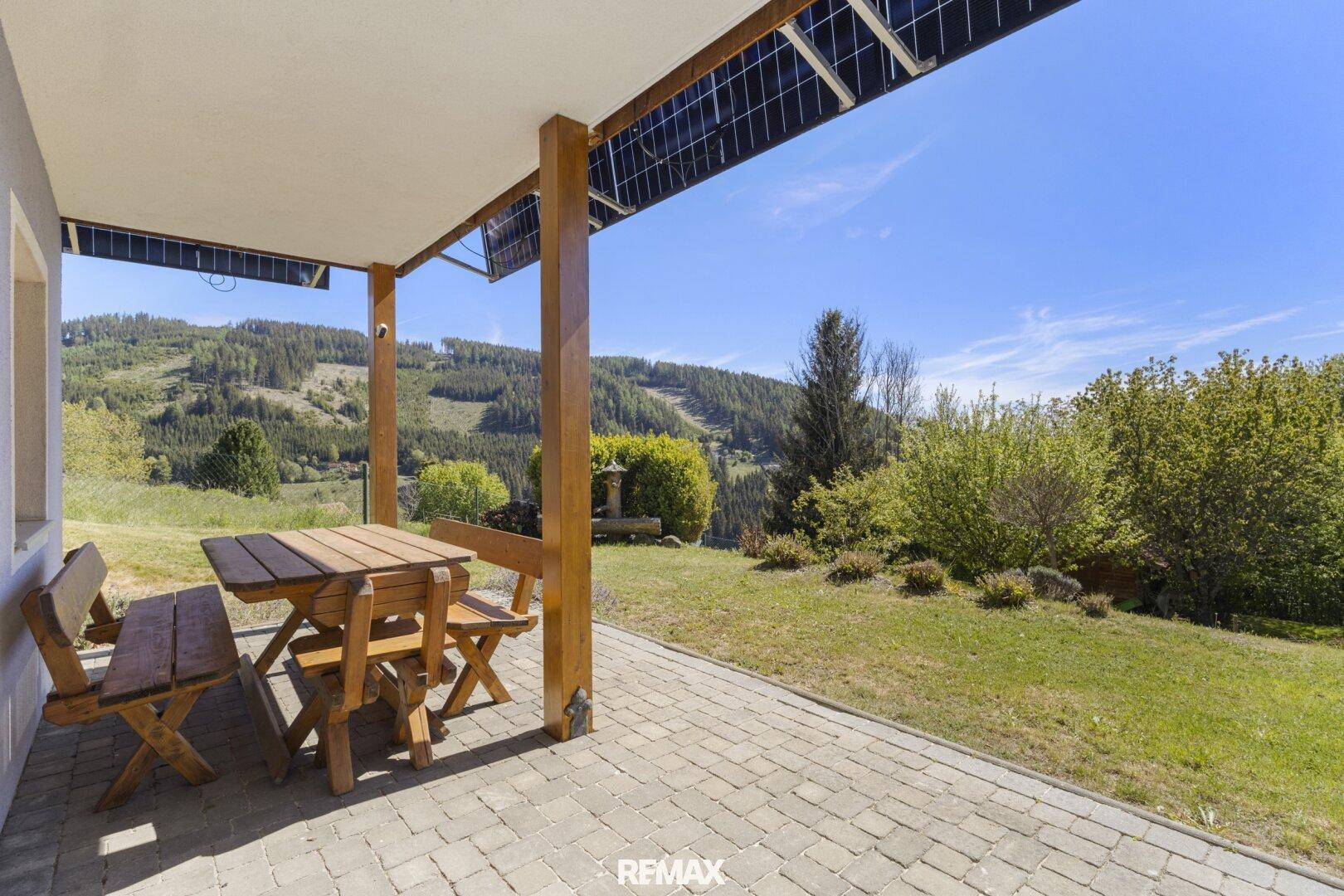 Überdachte Terrasse mit Holztisch und Bänken, umgeben von Natur und Bergblick.