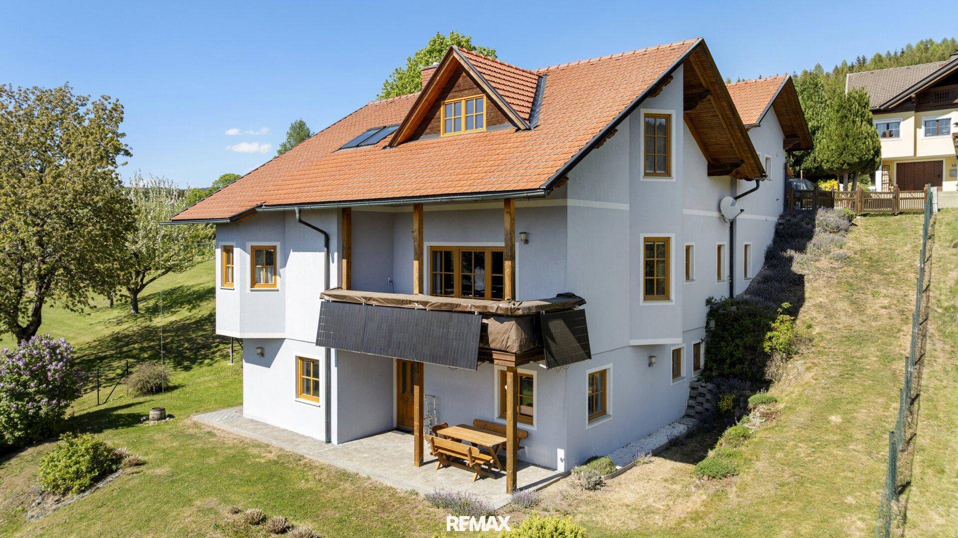 Vorderansicht des Hauses mit kleiner gepflasterter Terrasse und Holzbänken, umgeben von Grün.
