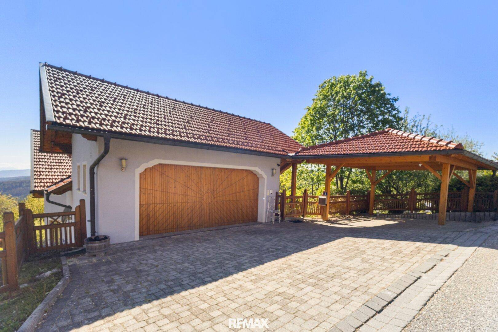 Doppelgarage mit Holztoren und angrenzendem Carport auf gepflasterter Zufahrt.