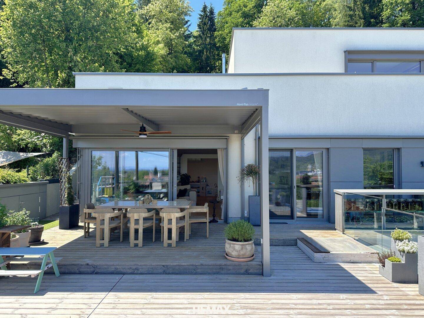 Ansicht des Hauses von der Terrasse mit überdachtem Essbereich und viel Grün.