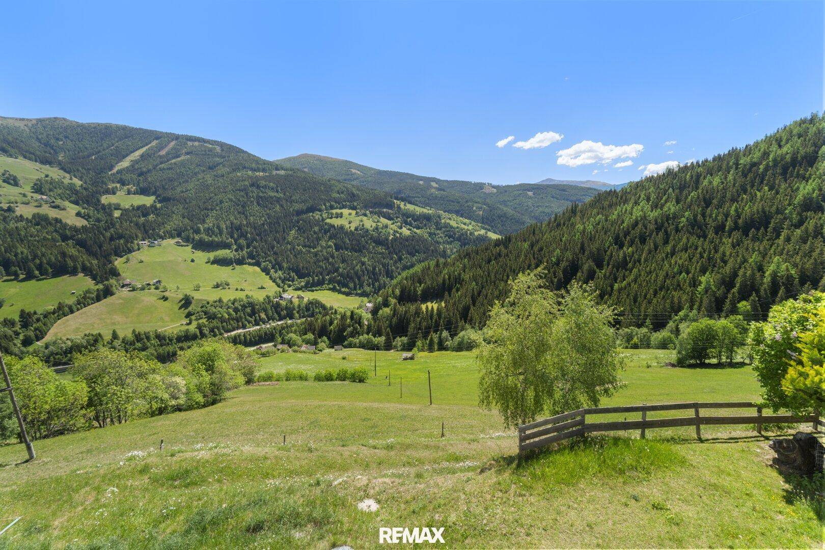 Weitläufiger Panoramablick über ein grünes Tal mit bewaldeten Bergen und Wiesen unter klarem Himmel.