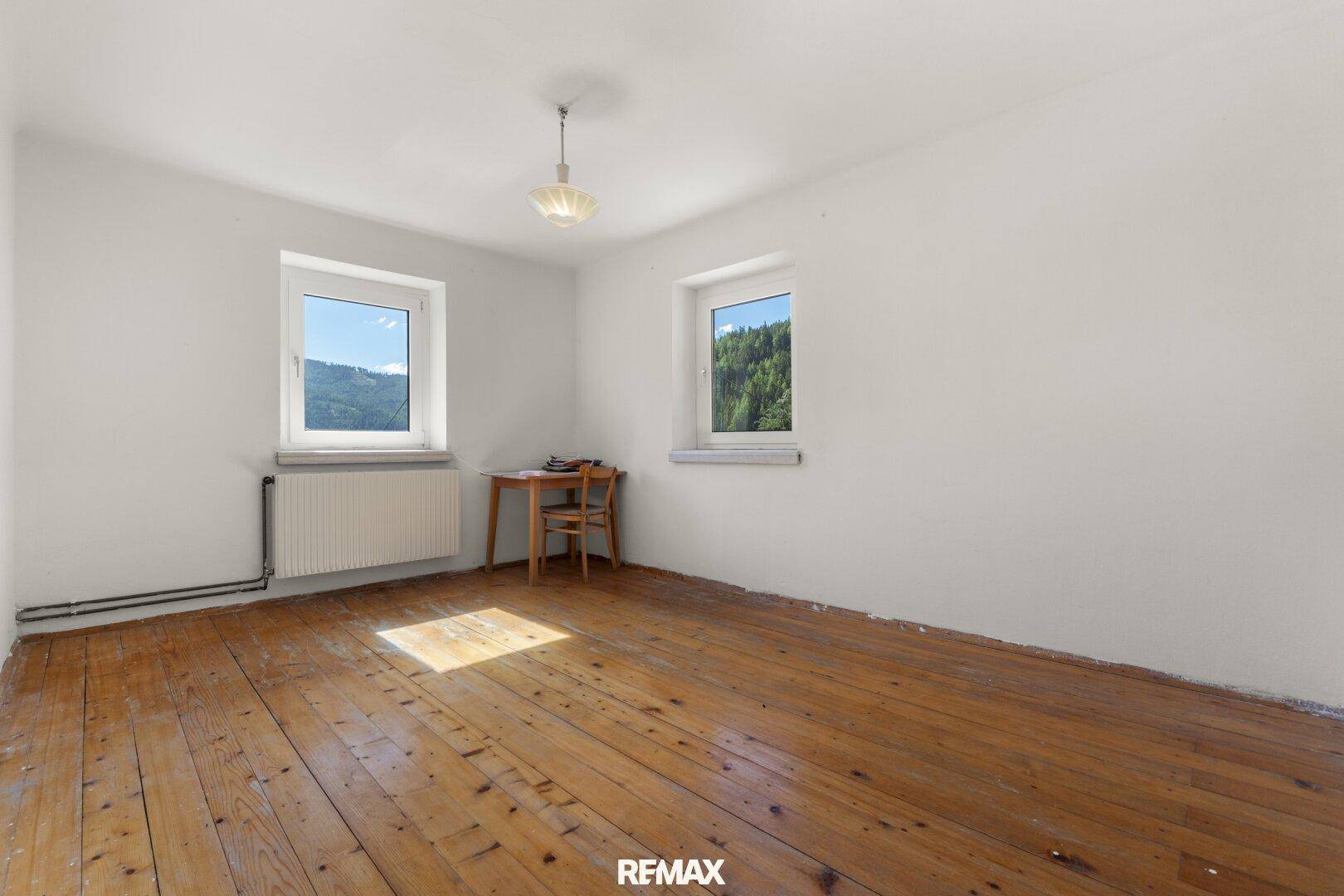 Leerer Raum mit weißen Wänden, originalem Holzdielenboden und zwei Fenstern mit Blick ins Grüne.