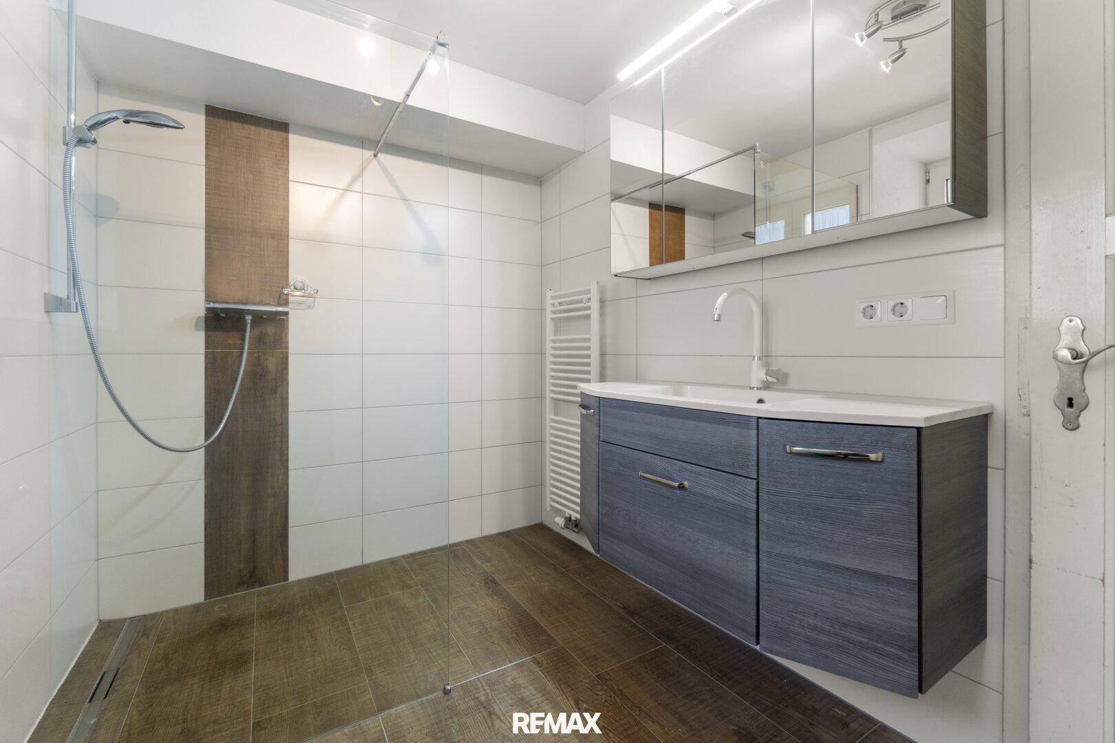 Modernes Badezimmer mit ebenerdiger Dusche, Waschtisch mit dunklem Unterschrank und weißen sowie Holzoptik-Fliesen.
