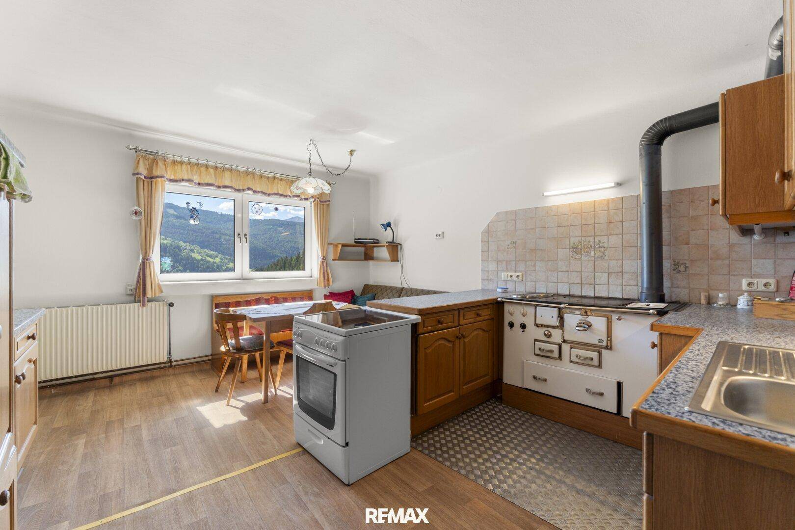 Geräumige Wohnküche mit traditionellem Holzofen, modernem Backofen und Esstisch am Fenster mit Bergblick.