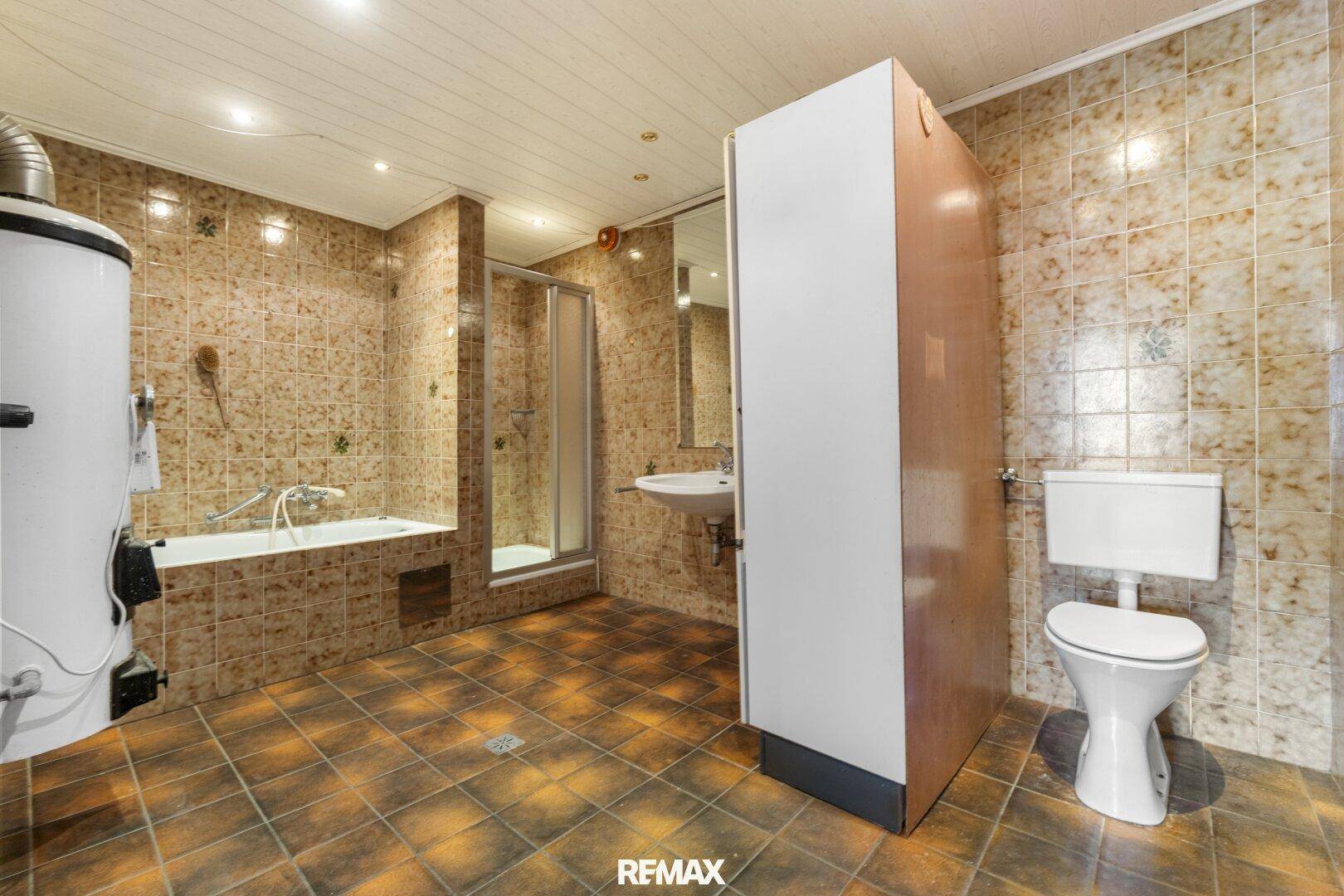 Großes, älteres Badezimmer mit gemusterten braunen Fliesen, Badewanne, Duschkabine, Toilette und Waschbecken.