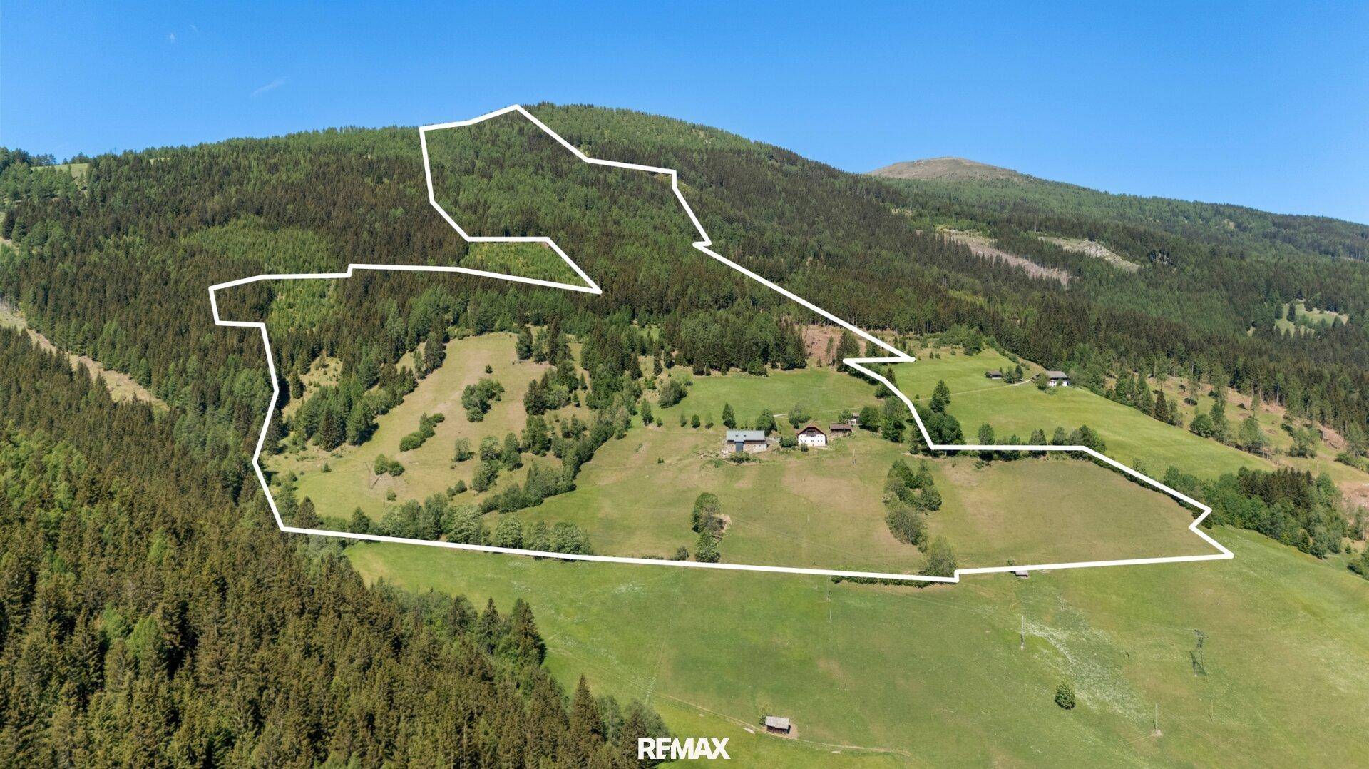 Luftaufnahme eines weitläufigen Berggebiets mit Wäldern und Wiesen, umrandet von einer Grundstücksgrenze.