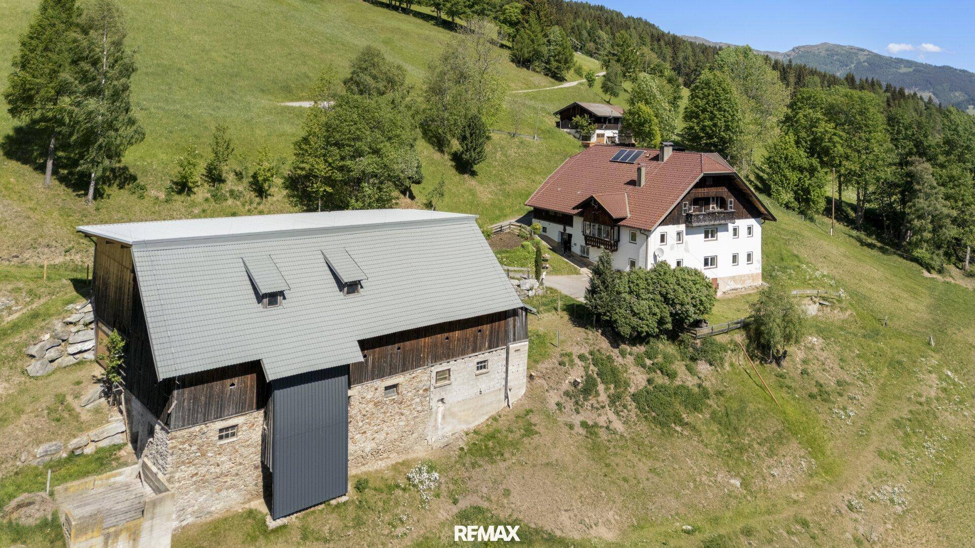 Luftaufnahme eines traditionellen Bauernhofs mit Wohnhaus und Scheune, eingebettet in eine grüne Hügellandschaft.