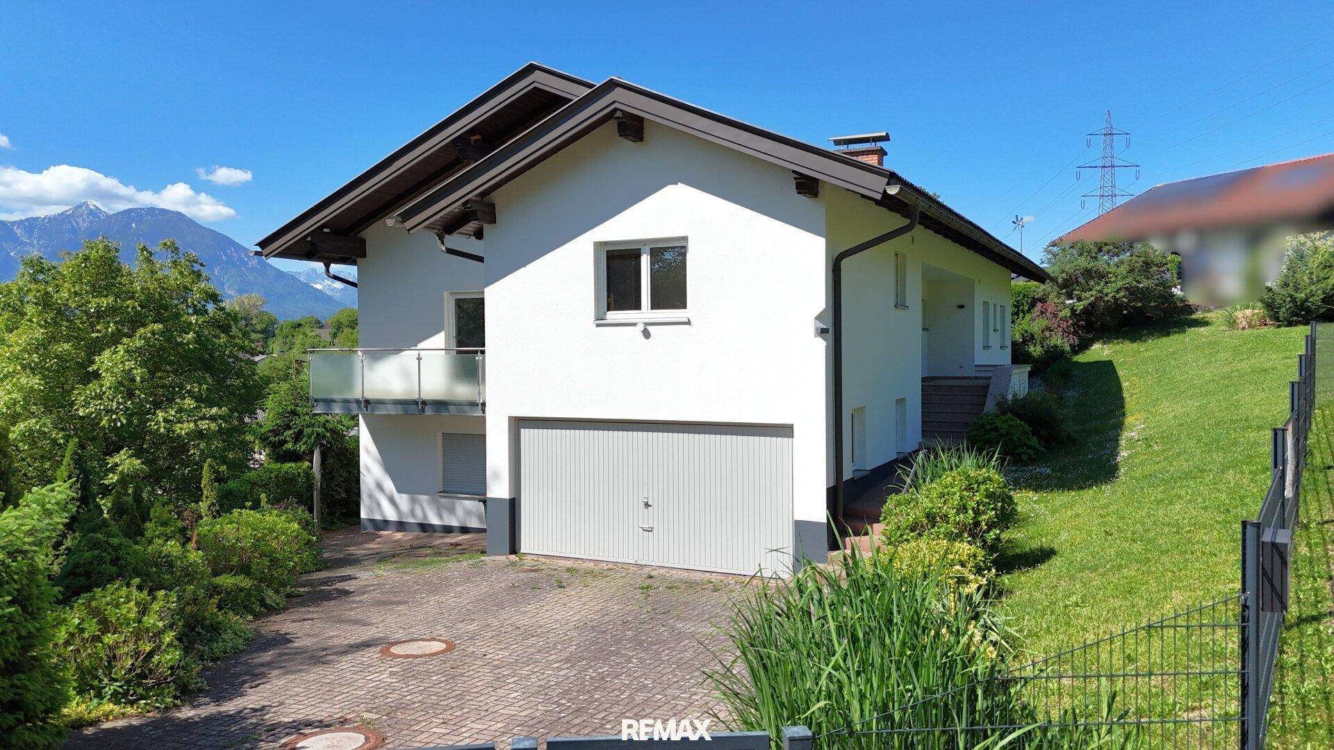 Vorderansicht des Hauses mit integrierter Garage und gepflasterter Auffahrt vor Bergkulisse.