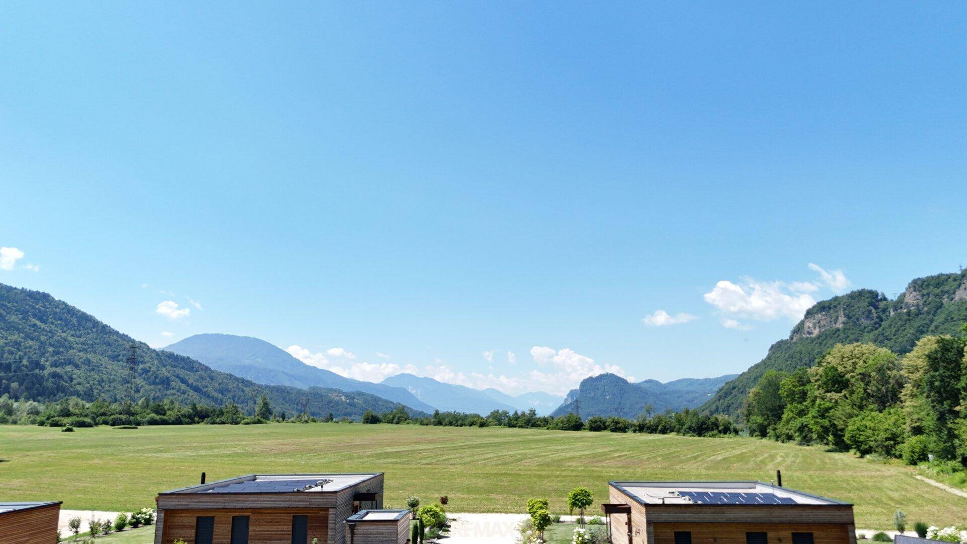 Weitläufige Landschaft mit grünen Feldern, modernen Häusern und majestätischen Bergen am Horizont.