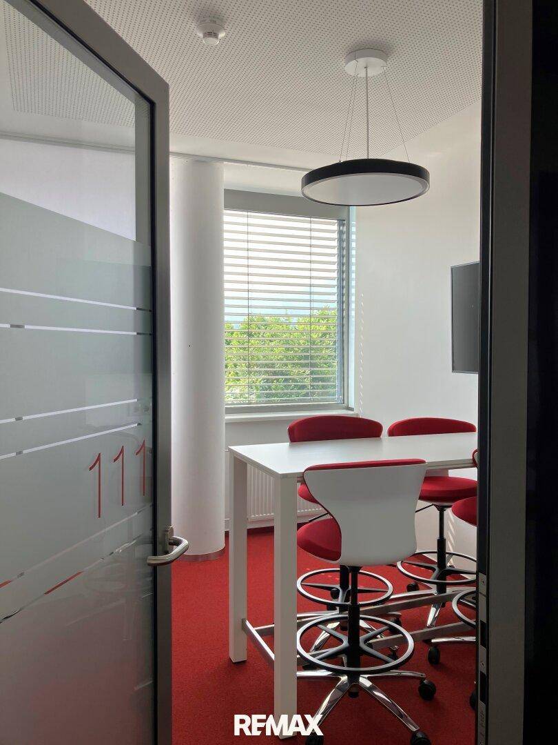 Moderner Besprechungsraum mit rotem Teppichboden und großem Fenster für natürliches Licht.