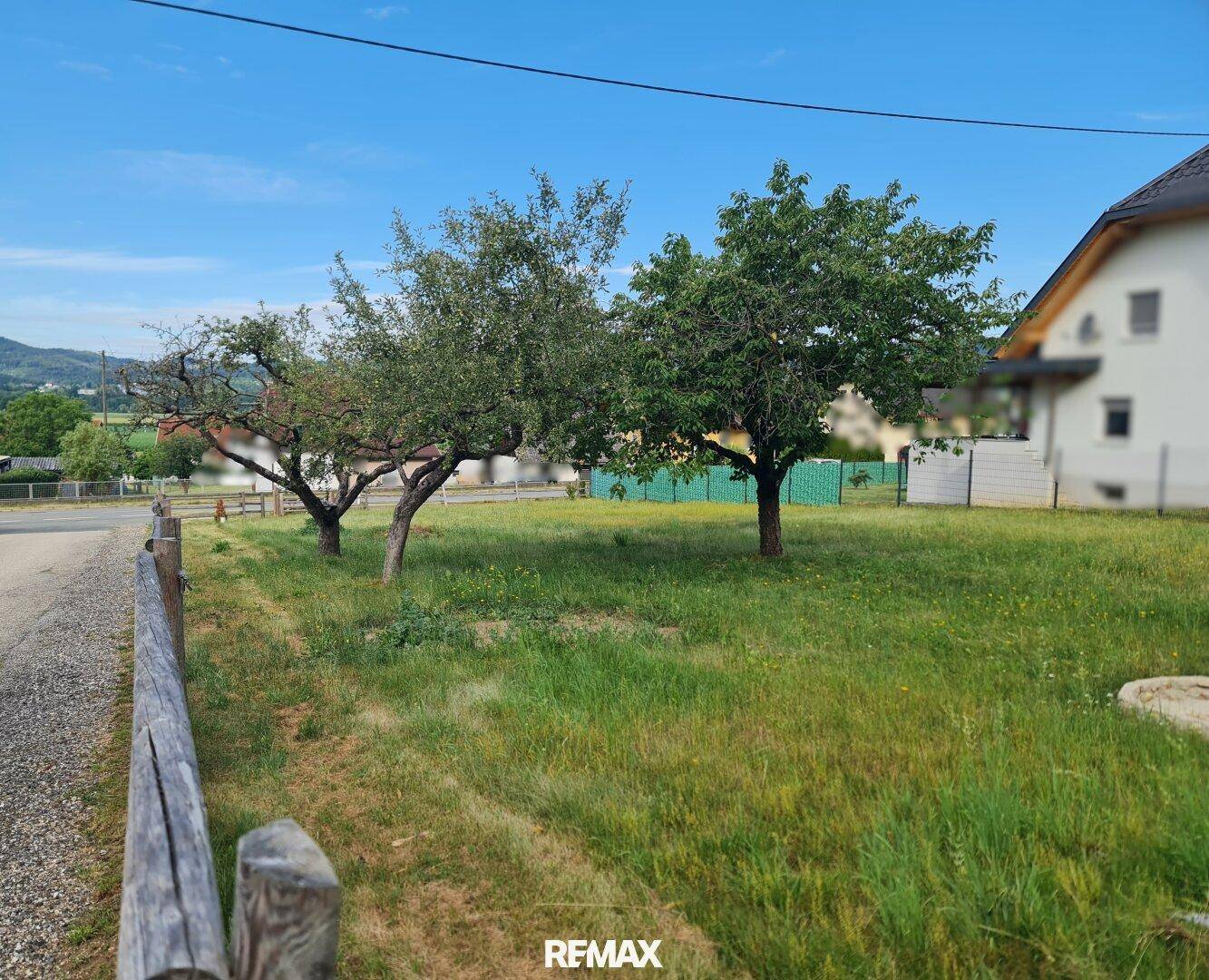 Grünes Grundstück mit Obstbäumen und Holzzaun, angrenzend an eine Straße mit Blick auf die Landschaft.