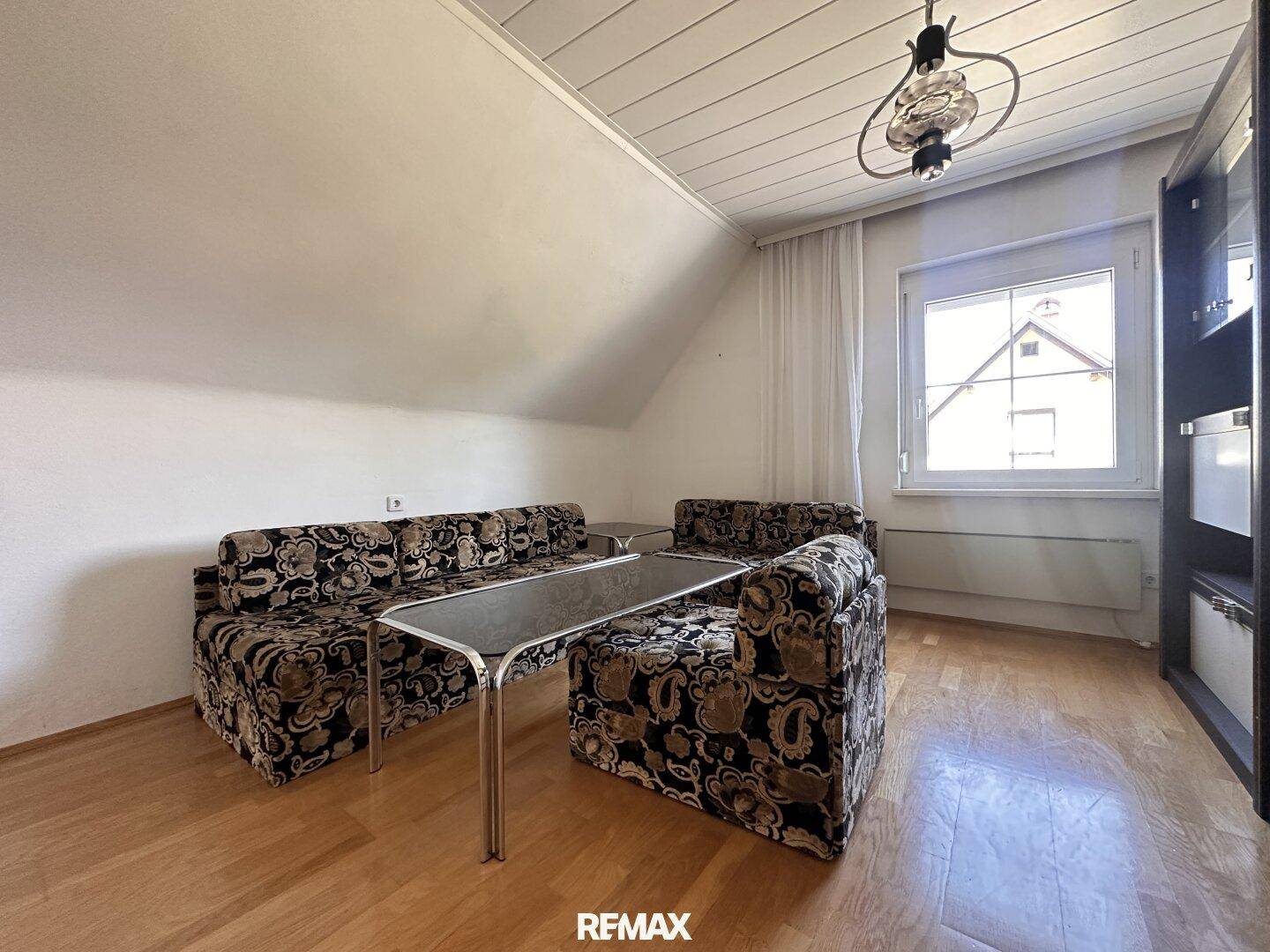 Gemütliches Zimmer im Dachgeschoss mit schrägen Wänden, Holzboden und einem Fenster mit Blick ins Grüne.