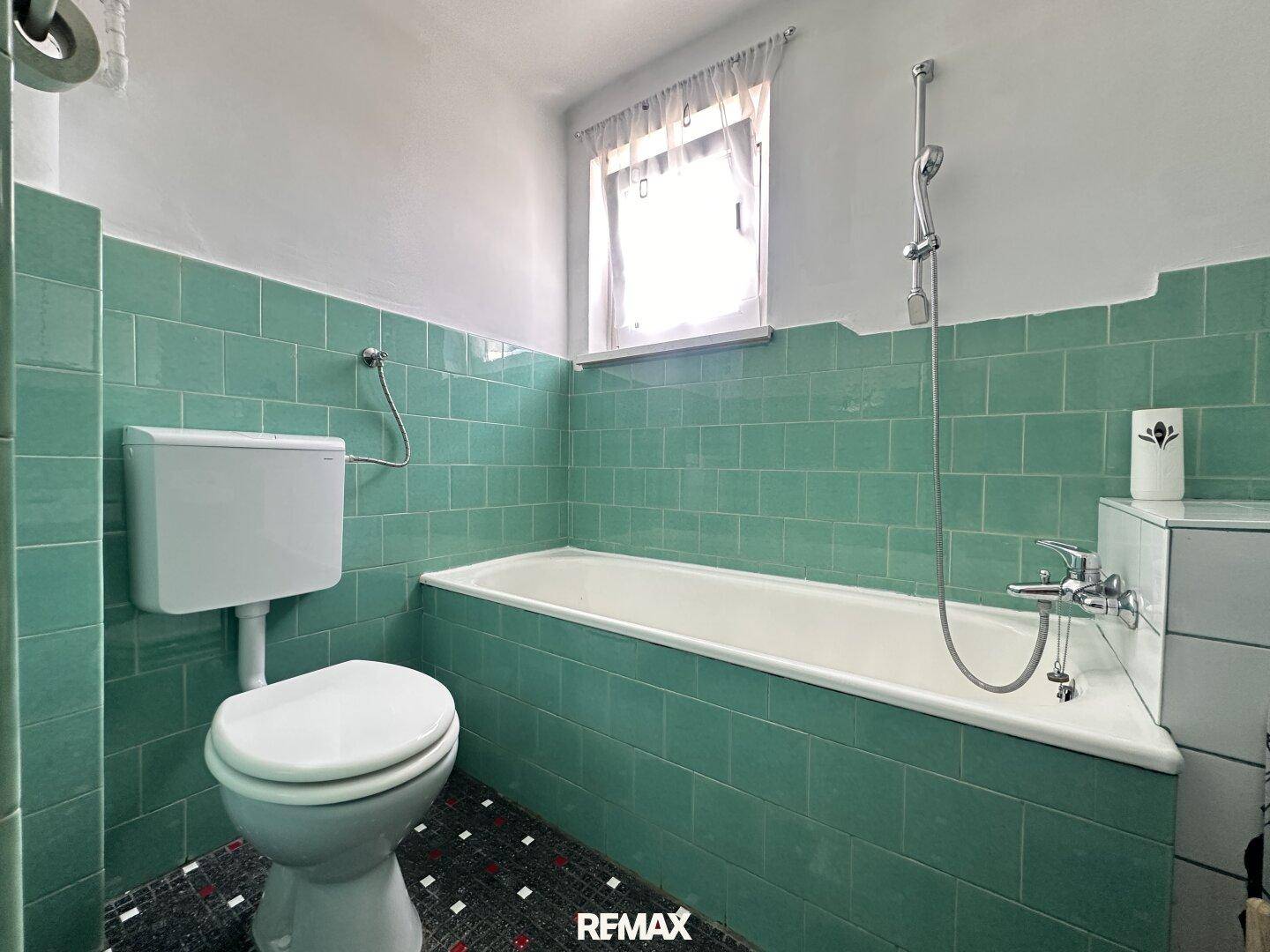 Badezimmer mit grünen Fliesen, Badewanne, Toilette und einem Fenster für natürliche Belüftung.