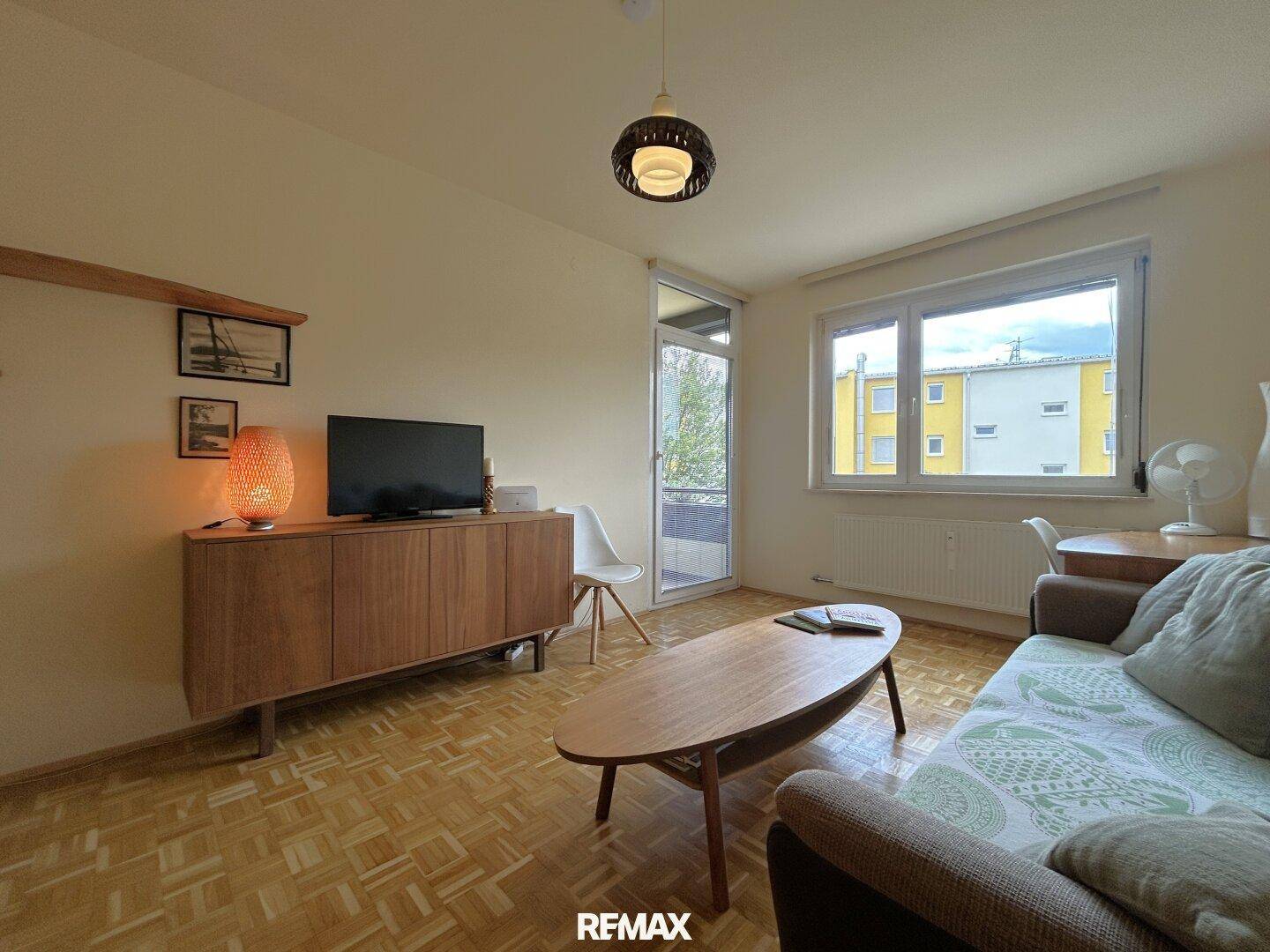 Wohnzimmer mit Parkettboden, TV-Möbel und großem Fenster mit Balkonzugang.