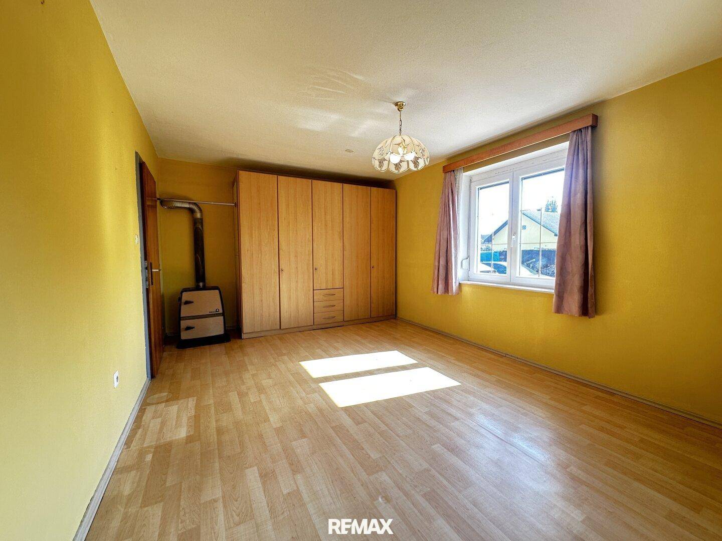 Helles Schlafzimmer mit gelben Wänden, Holzschrank, Fenster und einem gemütlichen Holzofen.