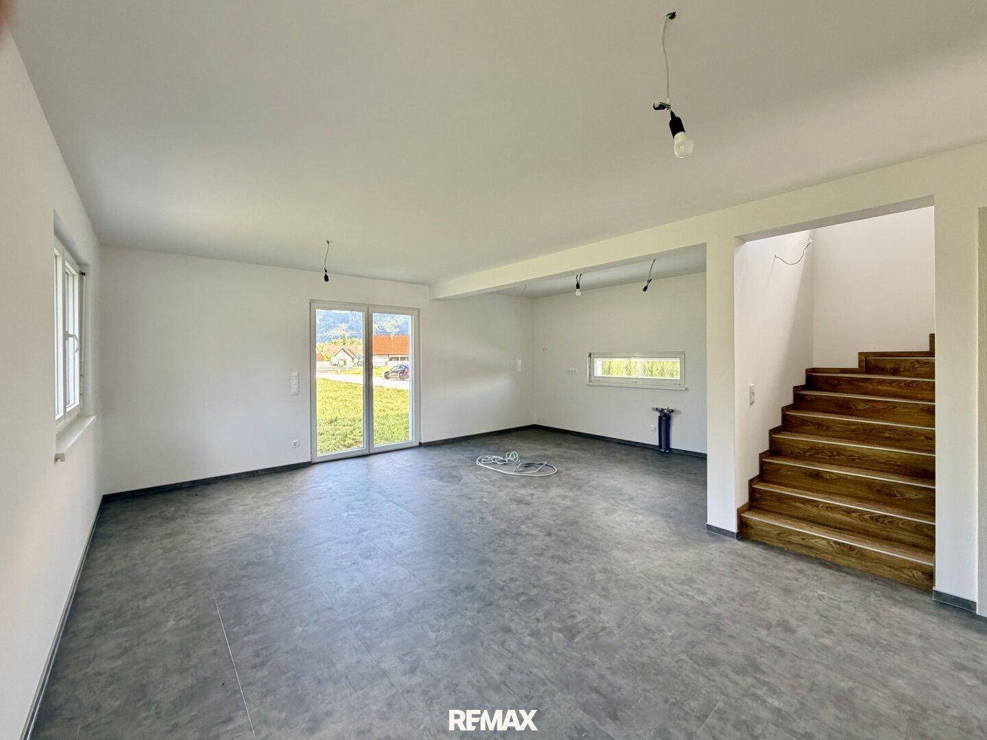 Geräumiger, unmöblierter Wohnbereich mit großen Fenstern und Zugang zum Garten, sowie einer Holztreppe.