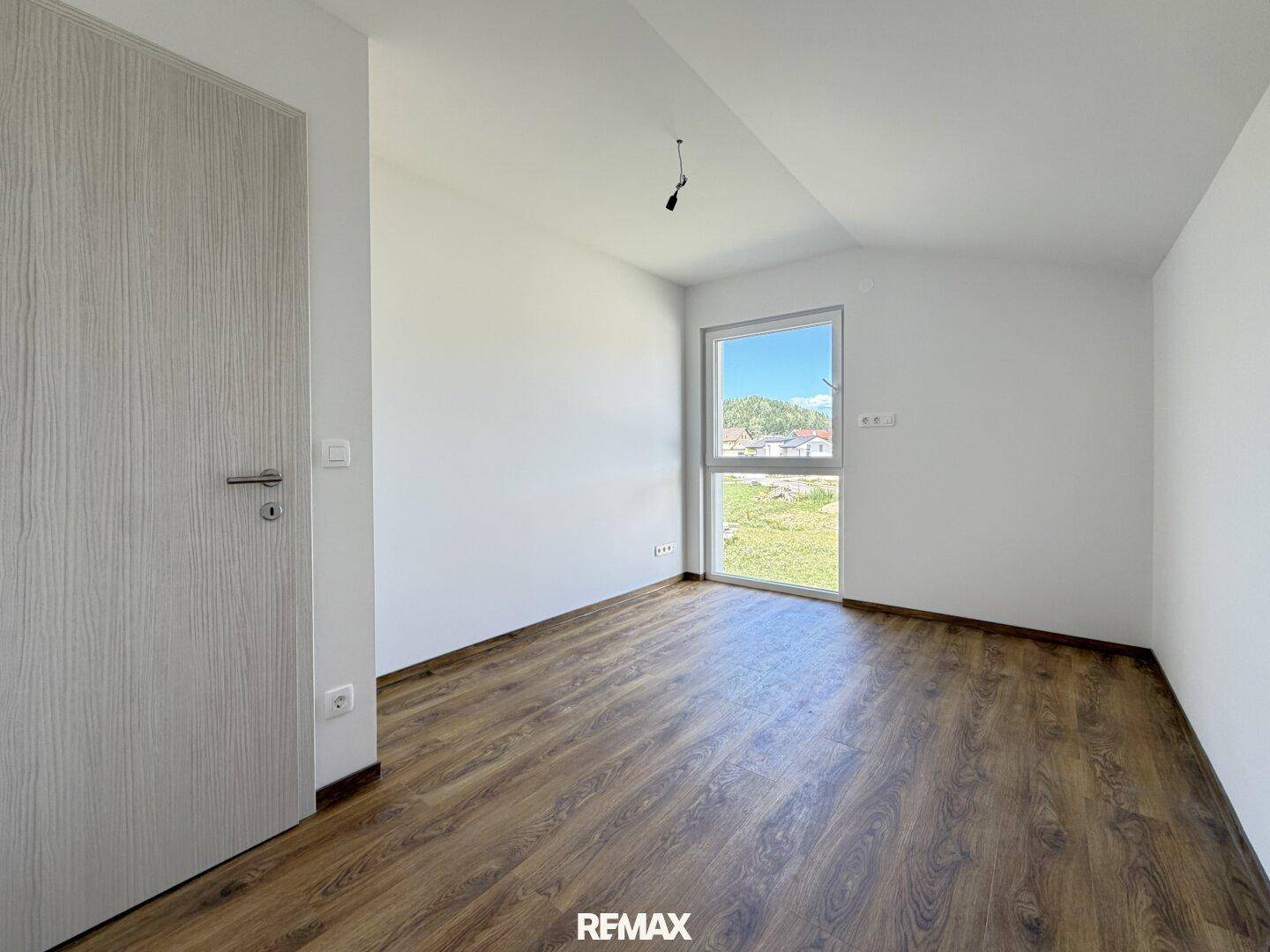 Helles, unmöbliertes Zimmer mit Holzboden und einem Fenster mit Blick ins Grüne.