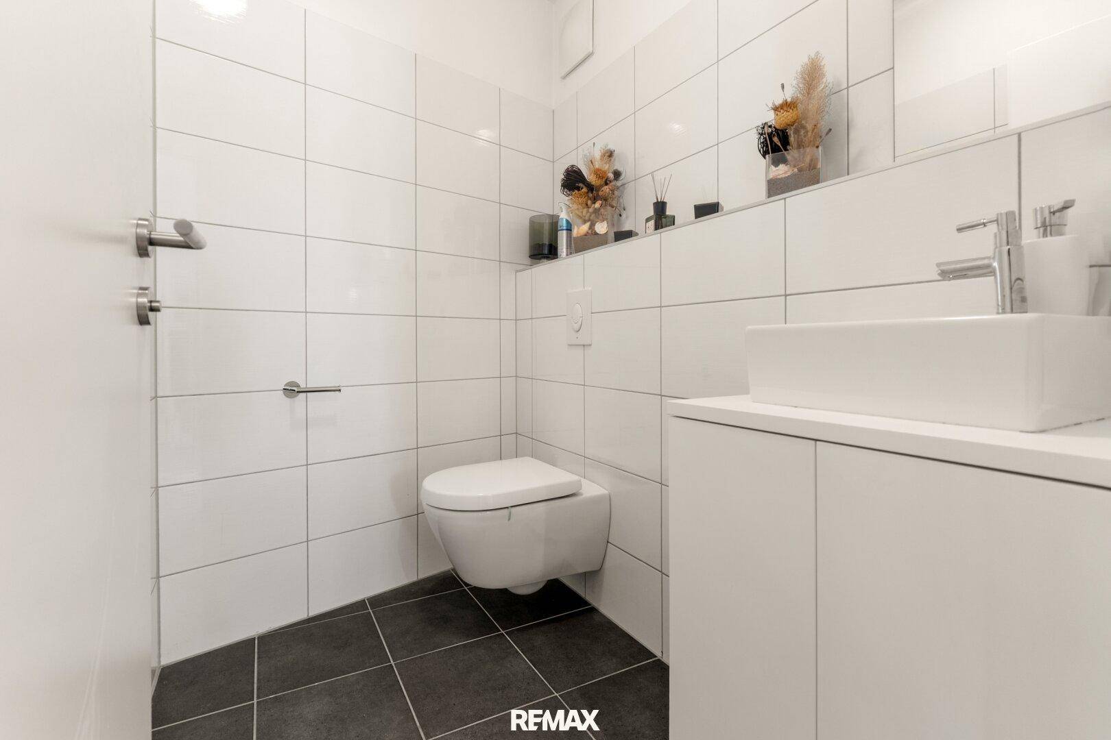 Helles Gäste-WC mit wandhängender Toilette, modernem Waschbecken und Fliesen in Weiß und Dunkelgrau.
