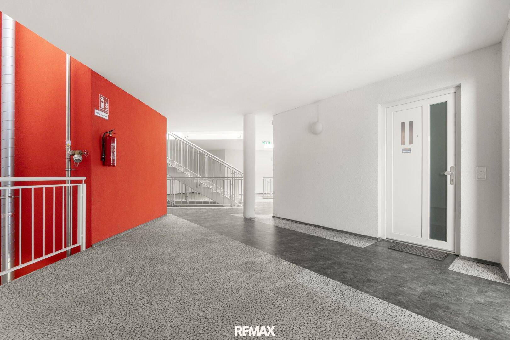 Heller Gemeinschaftsbereich im Gebäude mit roter Akzentwand und Treppenhaus im Hintergrund.