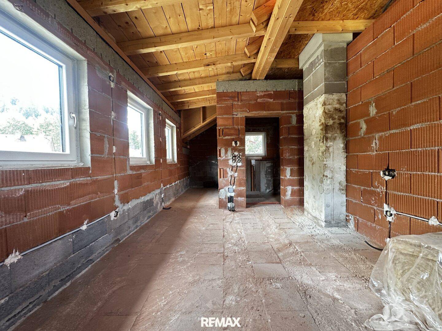 Raum im Rohbau mit Ziegelwänden, Holzdachstuhl und Fenstern im Dachgeschoss.