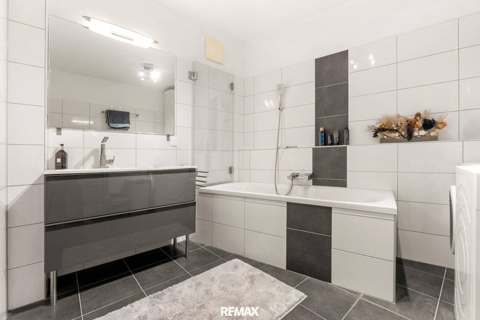 Modernes Badezimmer mit Badewanne, Dusche, Einzelwaschbecken und eleganten Fliesen in Weiß und Grau.