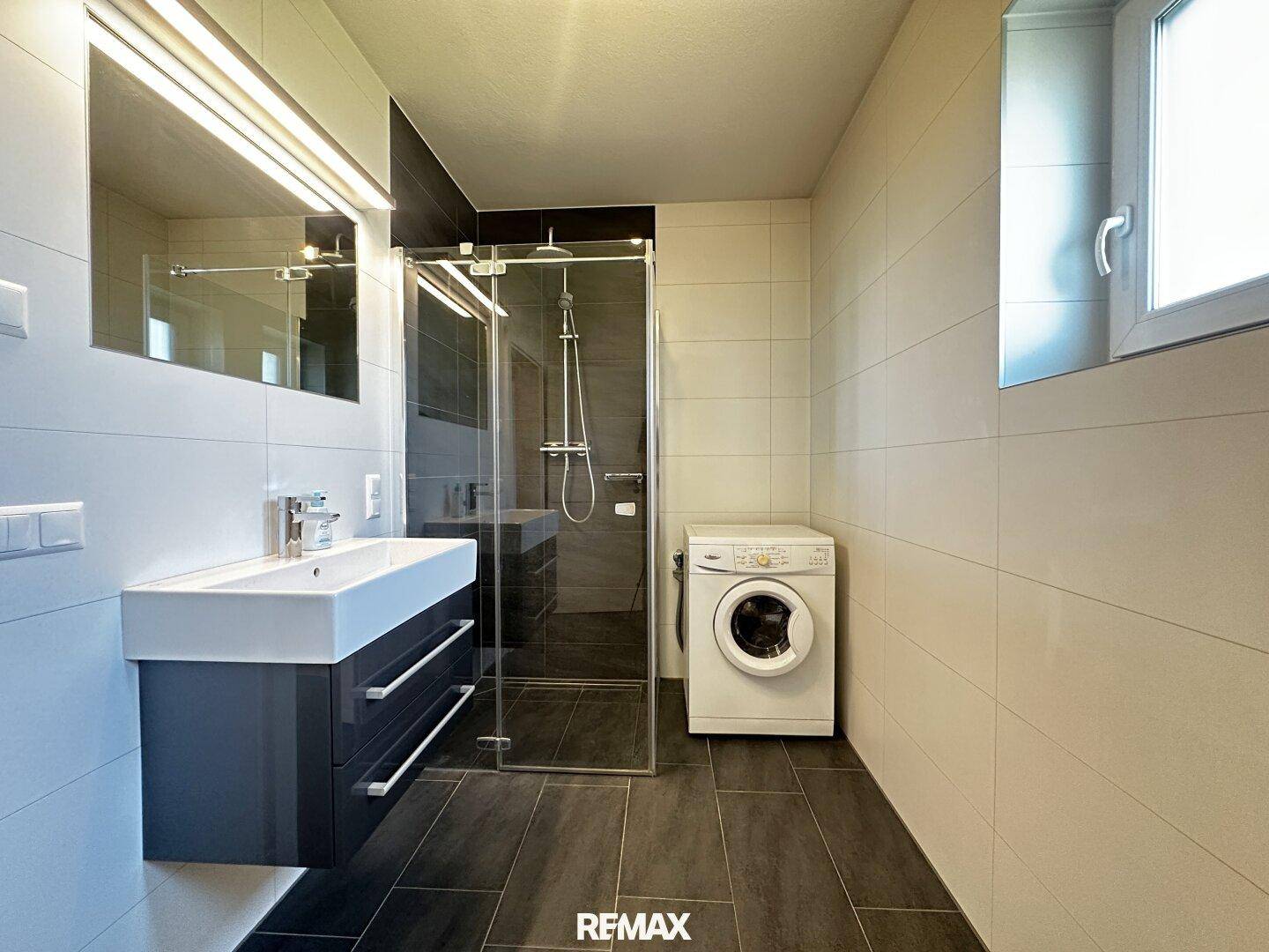 Modernes Badezimmer mit dunklen Fliesen, Duschkabine, Waschbecken und Waschmaschine.