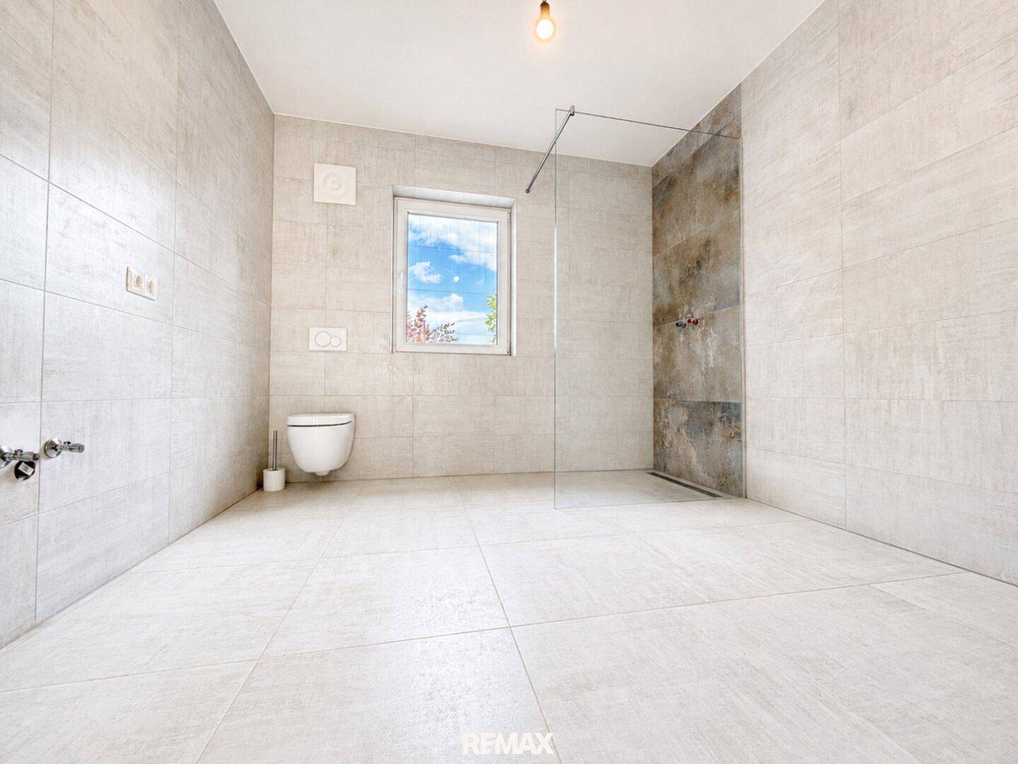 Helles, unmöbliertes Badezimmer mit ebenerdiger Dusche, Wand-WC und Fenster.