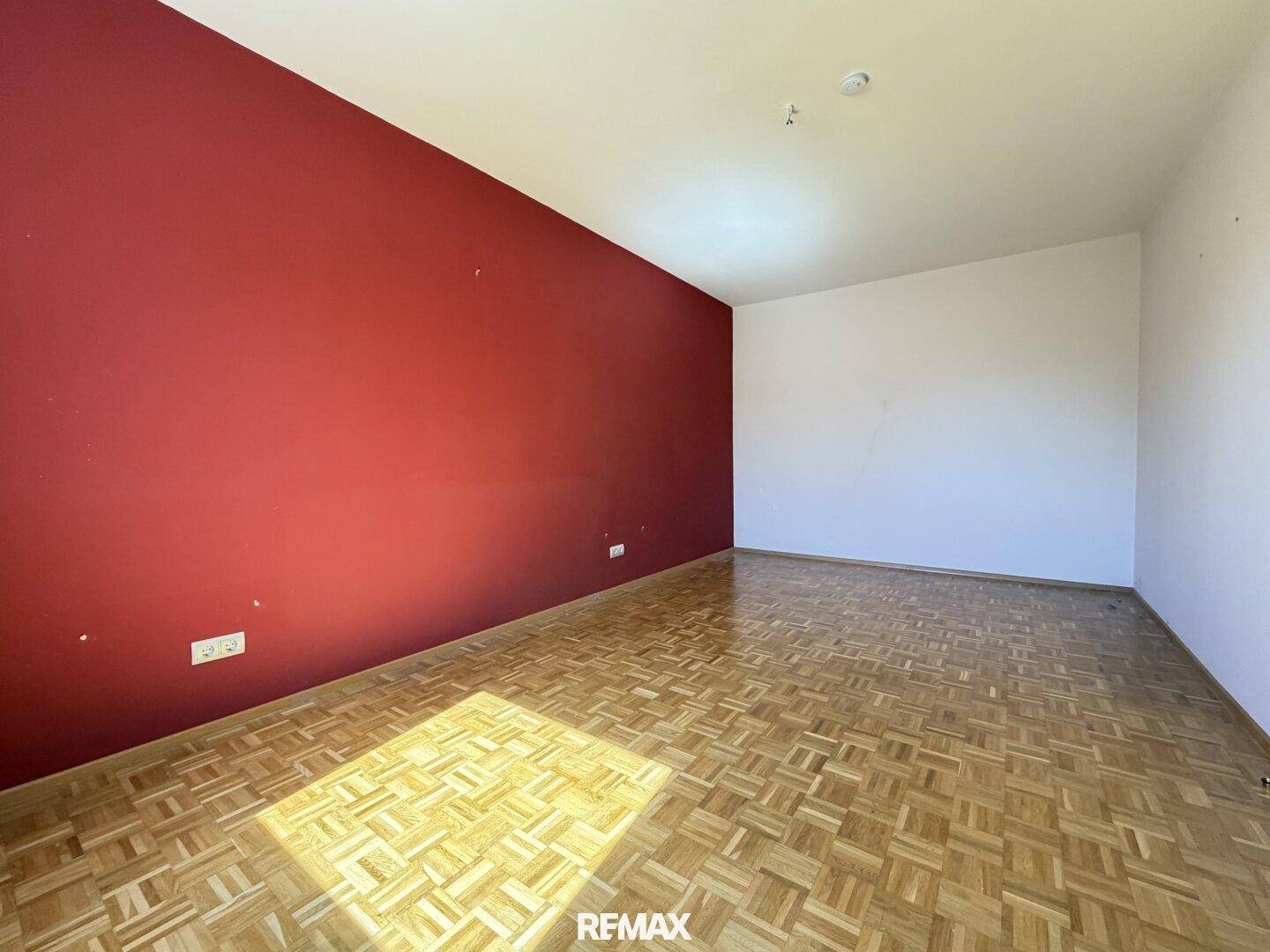 Helles Zimmer mit Parkettboden und einer auffälligen roten Akzentwand.