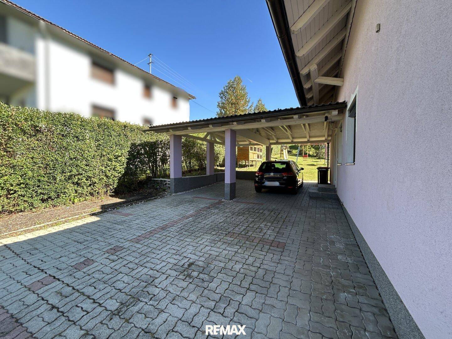 Überdachter Carport mit gepflasterter Einfahrt und grüner Hecke neben dem Wohnhaus.