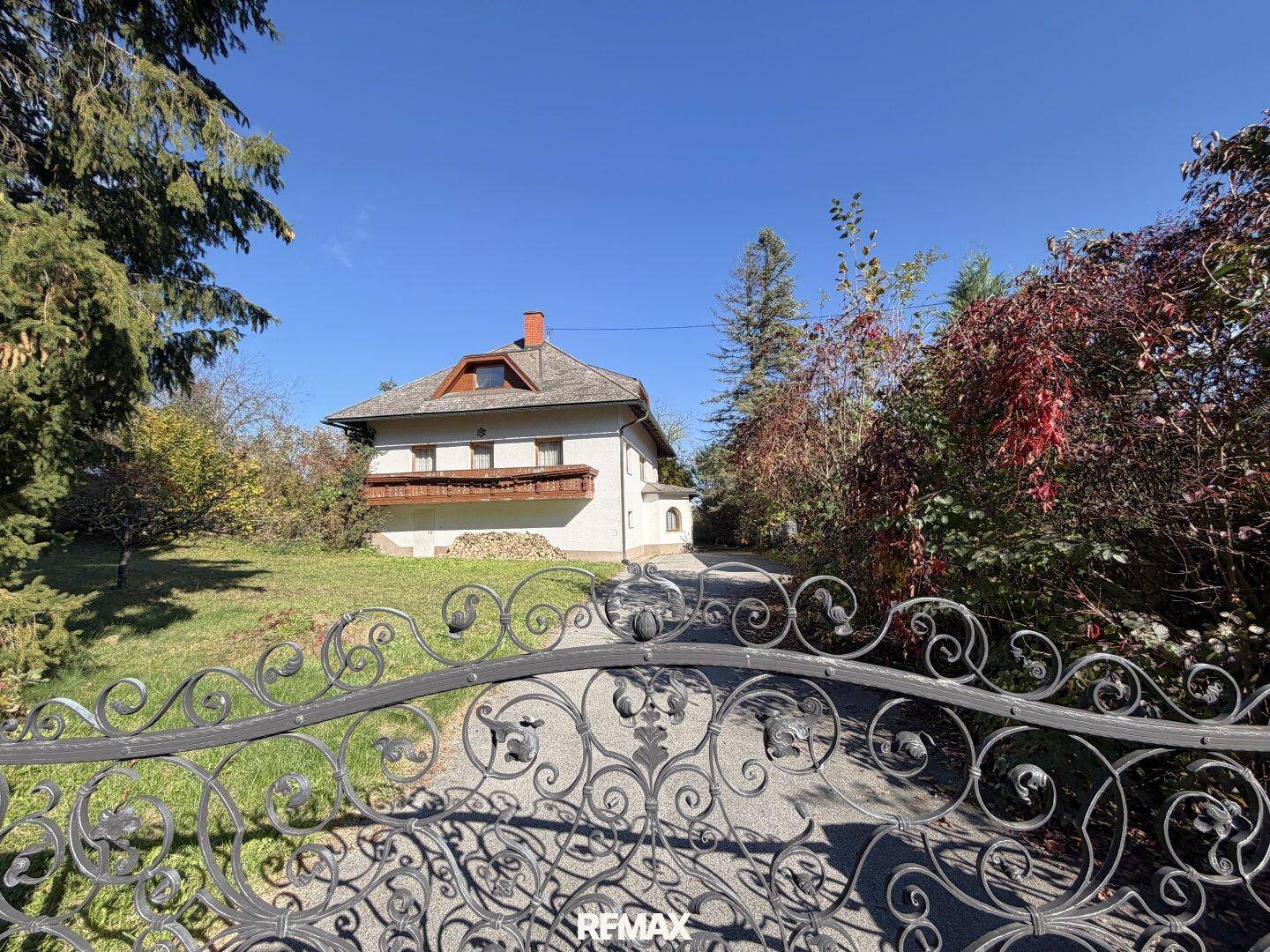 Ansicht des Hauses durch ein schmiedeeisernes Tor, umgeben von viel Grün und Bäumen.
