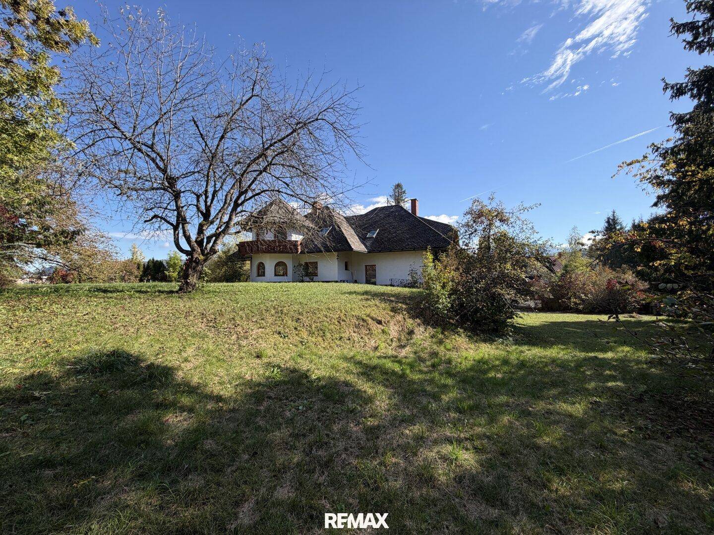 Weitläufiger Garten mit altem Baum und dem Haus im Hintergrund unter strahlend blauem Himmel.