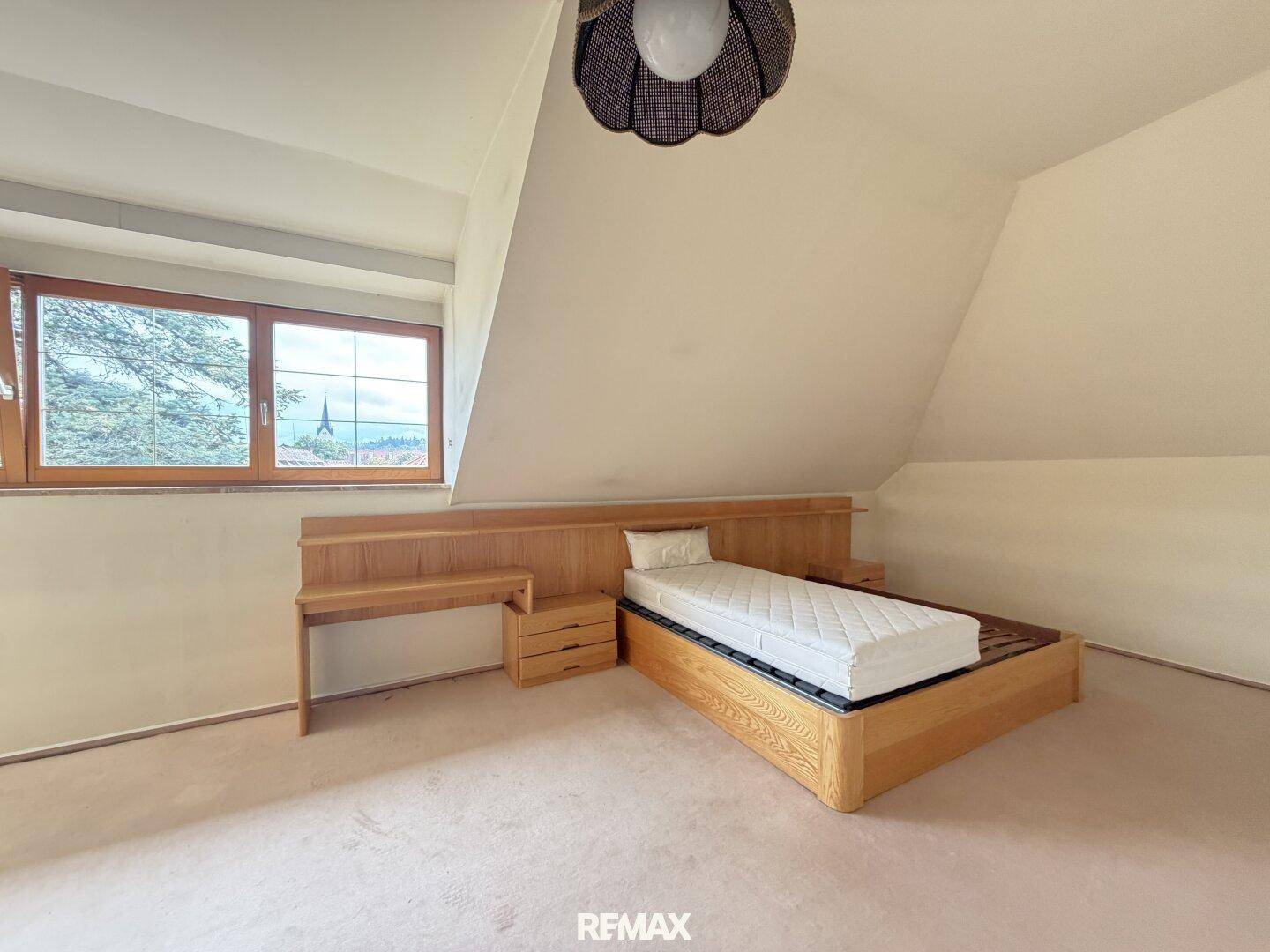 Gemütliches Schlafzimmer mit schräger Decke, Holzmöbeln und Fenster mit Blick ins Grüne.