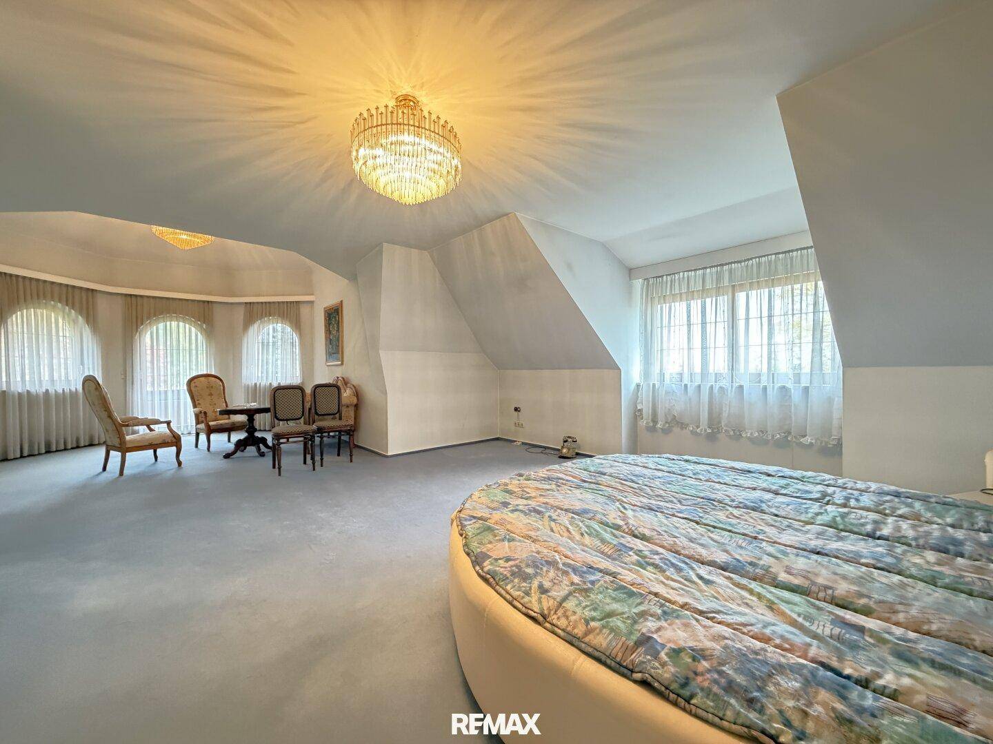 Großes Schlafzimmer im Dachgeschoss mit rundem Bett, Teppichboden und vielen Fenstern.
