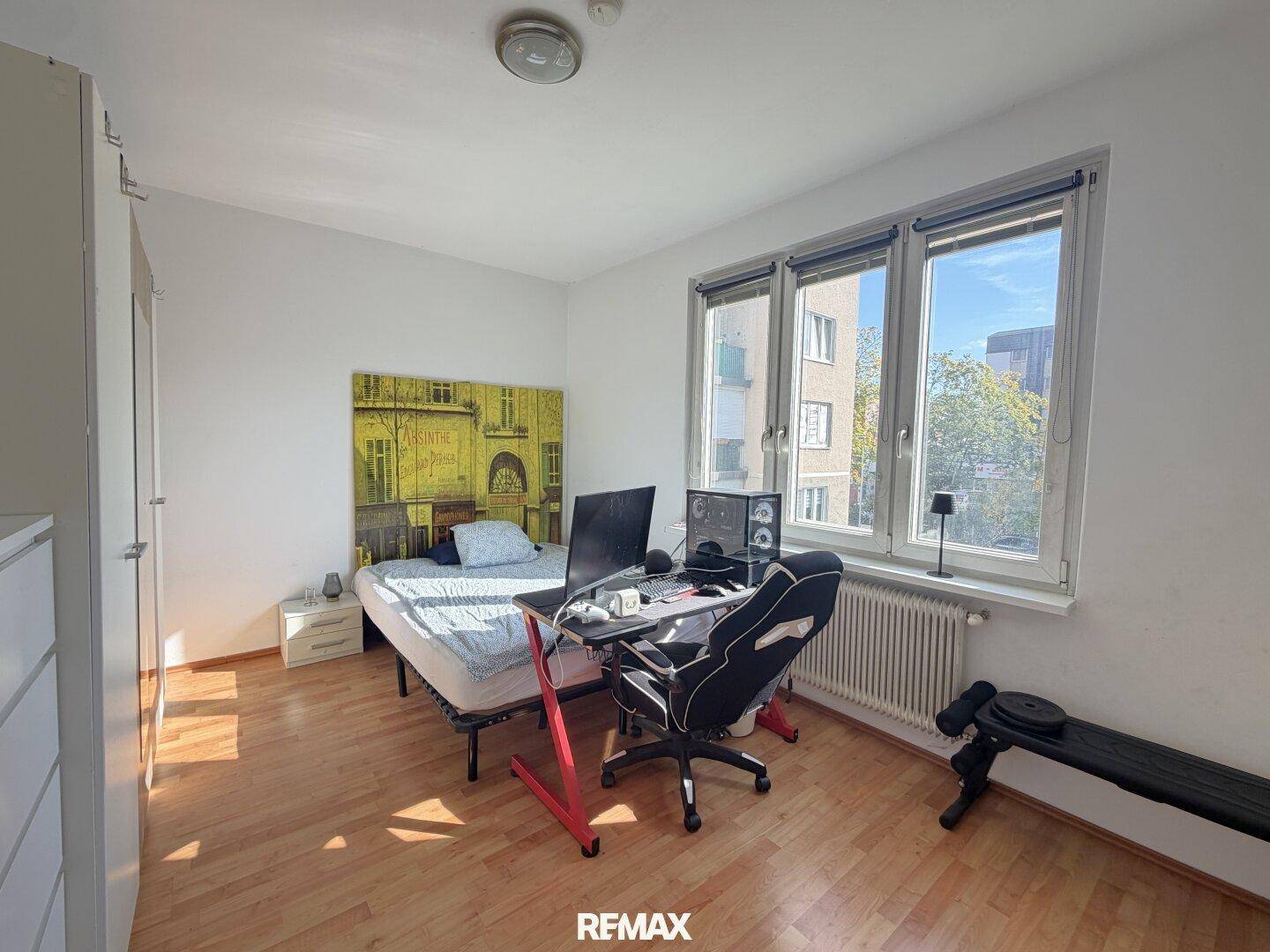 Schlafzimmer mit Einzelbett, Schreibtisch und einem großen Fenster, das den Raum hell erscheinen lässt.