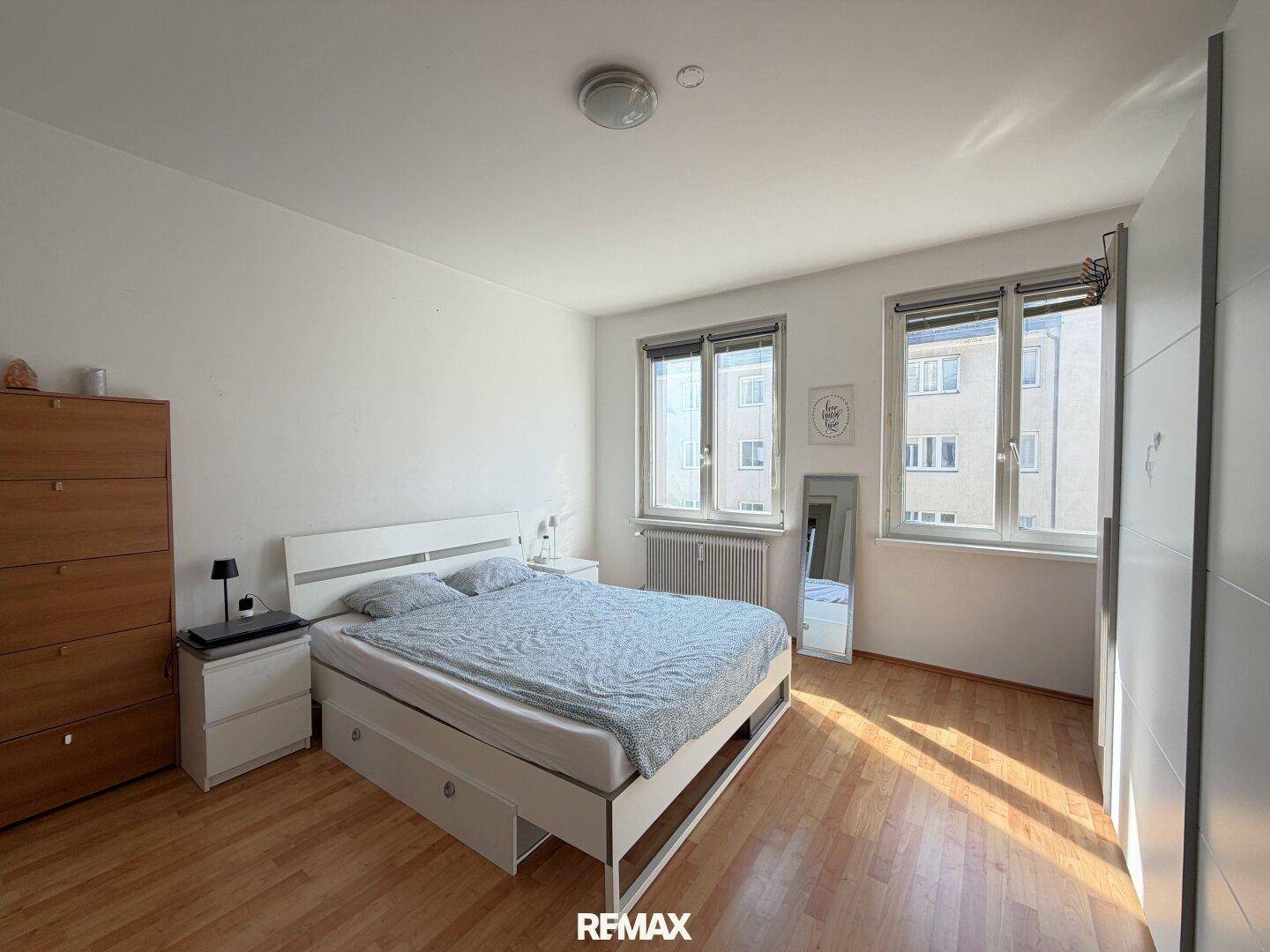 Helles Schlafzimmer mit Doppelbett, Holzboden und zwei Fenstern, die den Raum gut belichten.