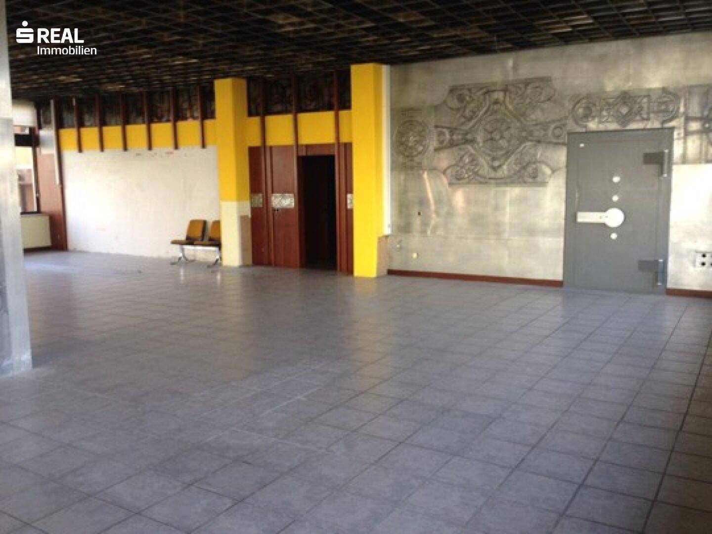 Geräumiger Gewerberaum mit Fliesenboden, gelben Säulen und einer großen Tresortür an der Wand.