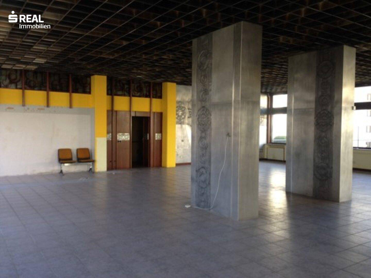 Weitläufiger Gewerberaum mit Fliesenboden, hohen Decken und markanten Betonpfeilern in der Mitte.