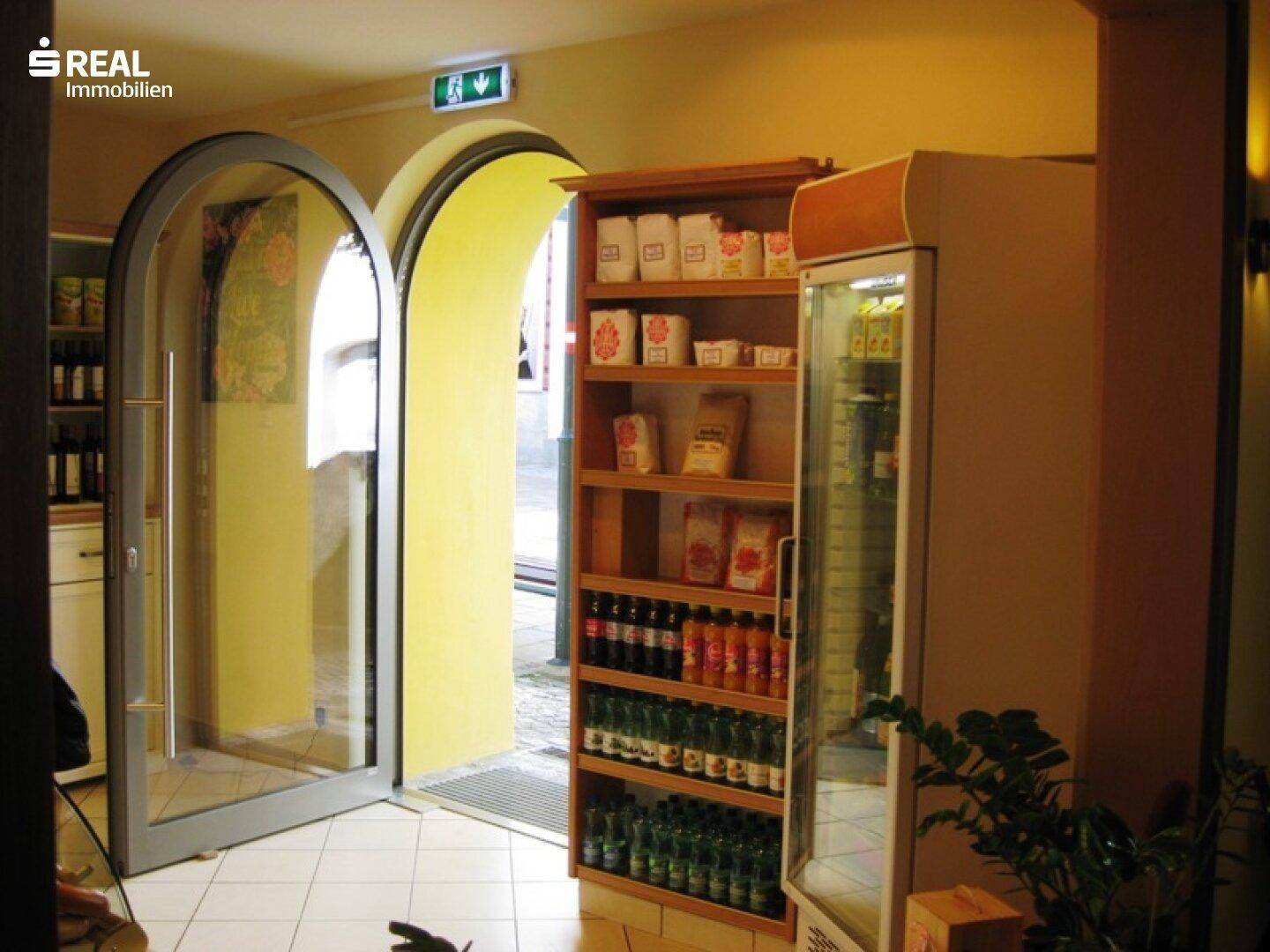 Eingangsbereich eines Geschäfts mit doppelten Glastüren, Regalen und einem Kühlschrank.