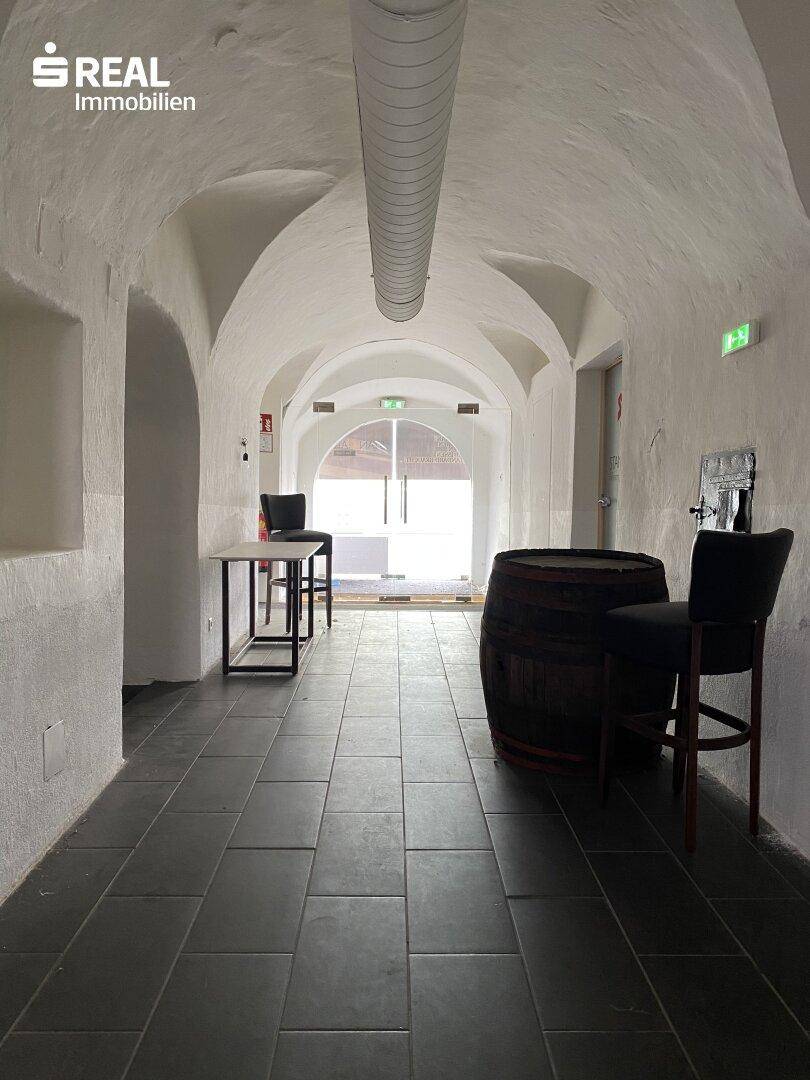 Langer Gewölbegang mit dunklen Fliesen, einem dekorativen Weinfass und moderner Beleuchtung am Ende.