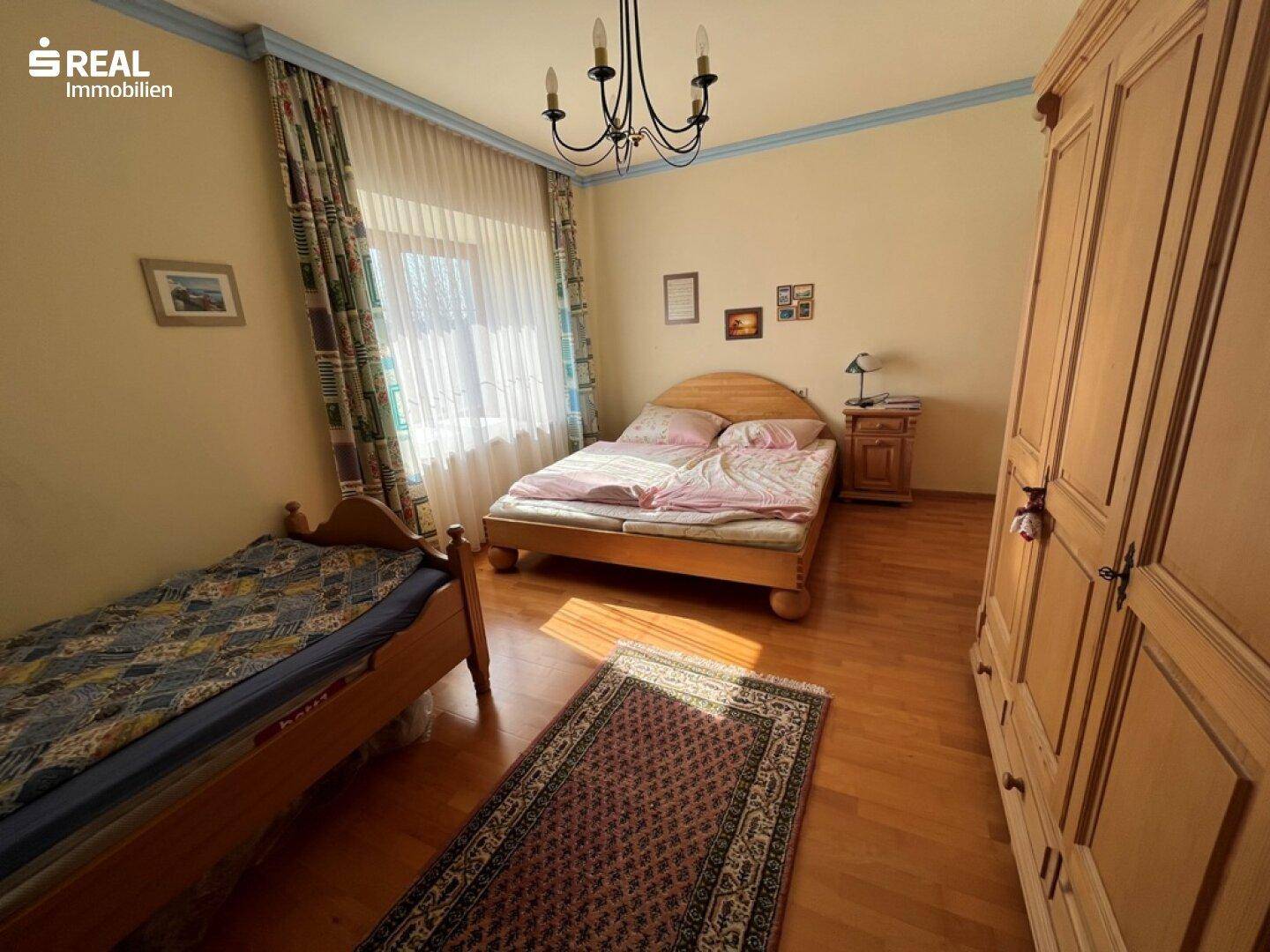 Helles Schlafzimmer mit zwei Holzbetten, einem großen Kleiderschrank und einem Fenster mit Vorhängen.