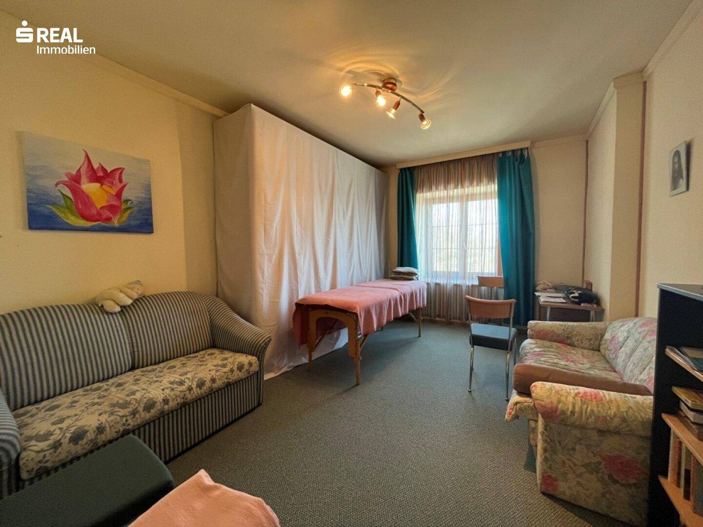Vielseitiger Raum mit zwei Sofas, einer Massageliege und einem Fenster mit blauen Vorhängen und Teppichboden.
