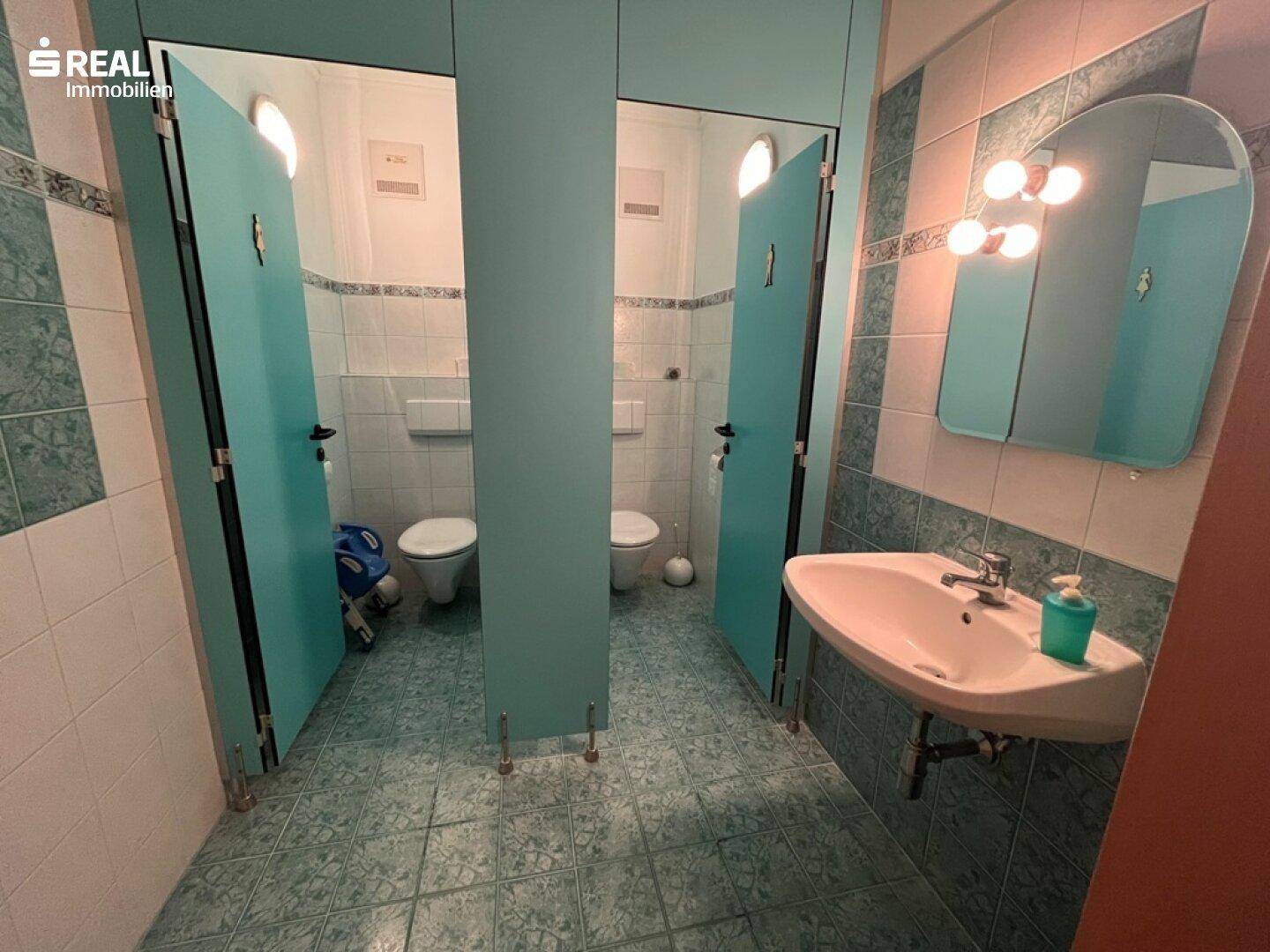 Sanitärbereich mit mehreren Toilettenkabinen, einem Waschbecken und blau-weiß gefliesten Wänden und Boden.