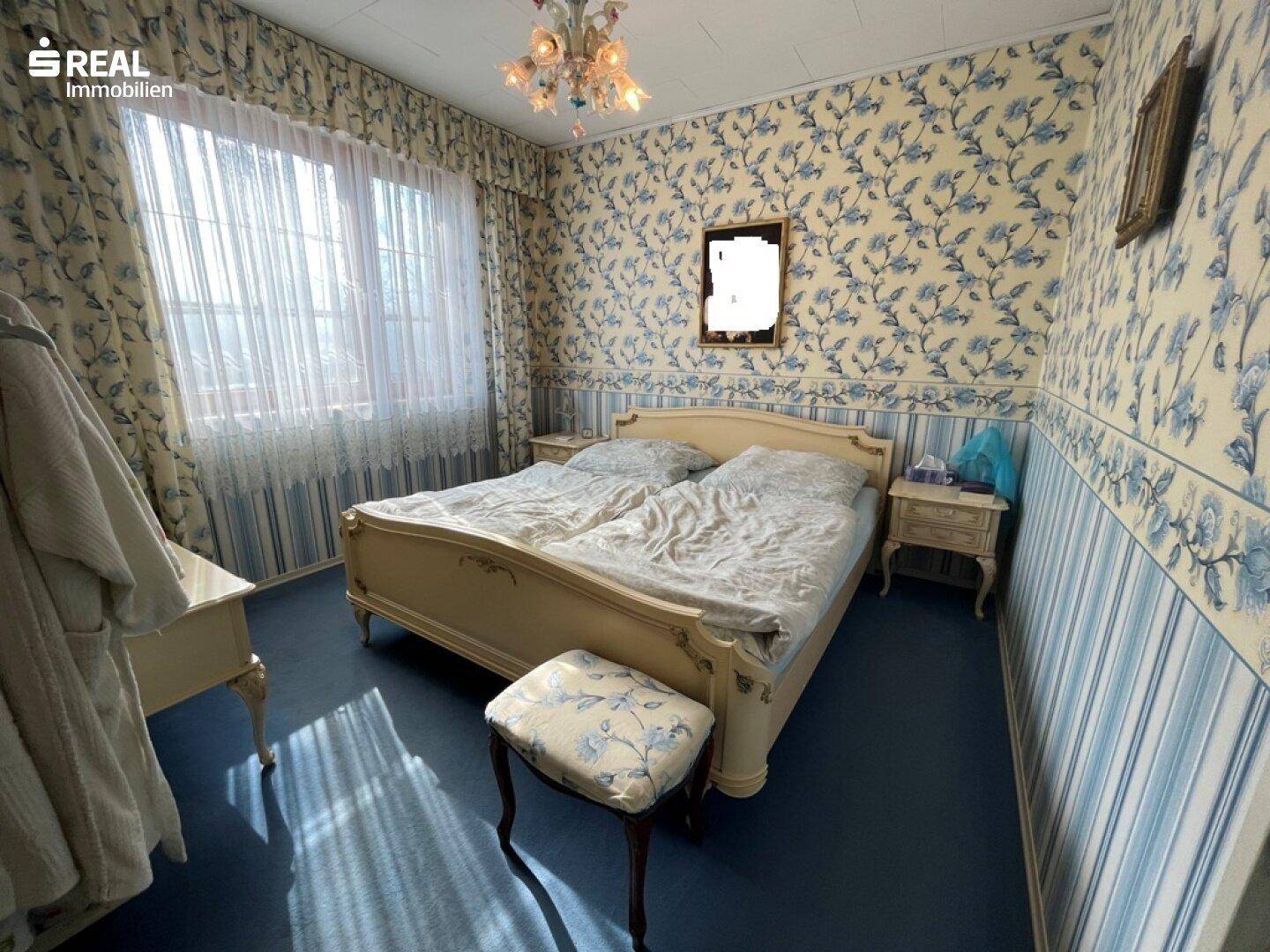 Schlafzimmer mit floraler Tapete, großem Bett, Nachttischen und einem Fenster mit Vorhängen und Kronleuchter.