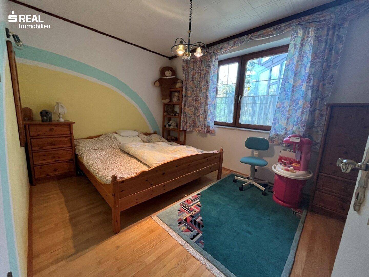 Kinderzimmer mit Holzbett, Kommode, Schreibtischstuhl und einer farbenfroh gestalteten Wand.