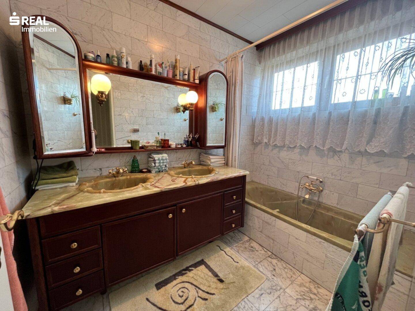 Badezimmer mit Doppelwaschbecken, Badewanne und Marmorfliesen an Wänden und Boden, sowie einem Fenster.