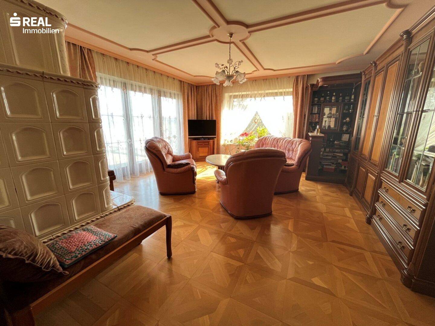 Elegantes Wohnzimmer mit Kachelofen, bequemen Sesseln, Parkettboden und großen Fenstern mit Stuckdecke.