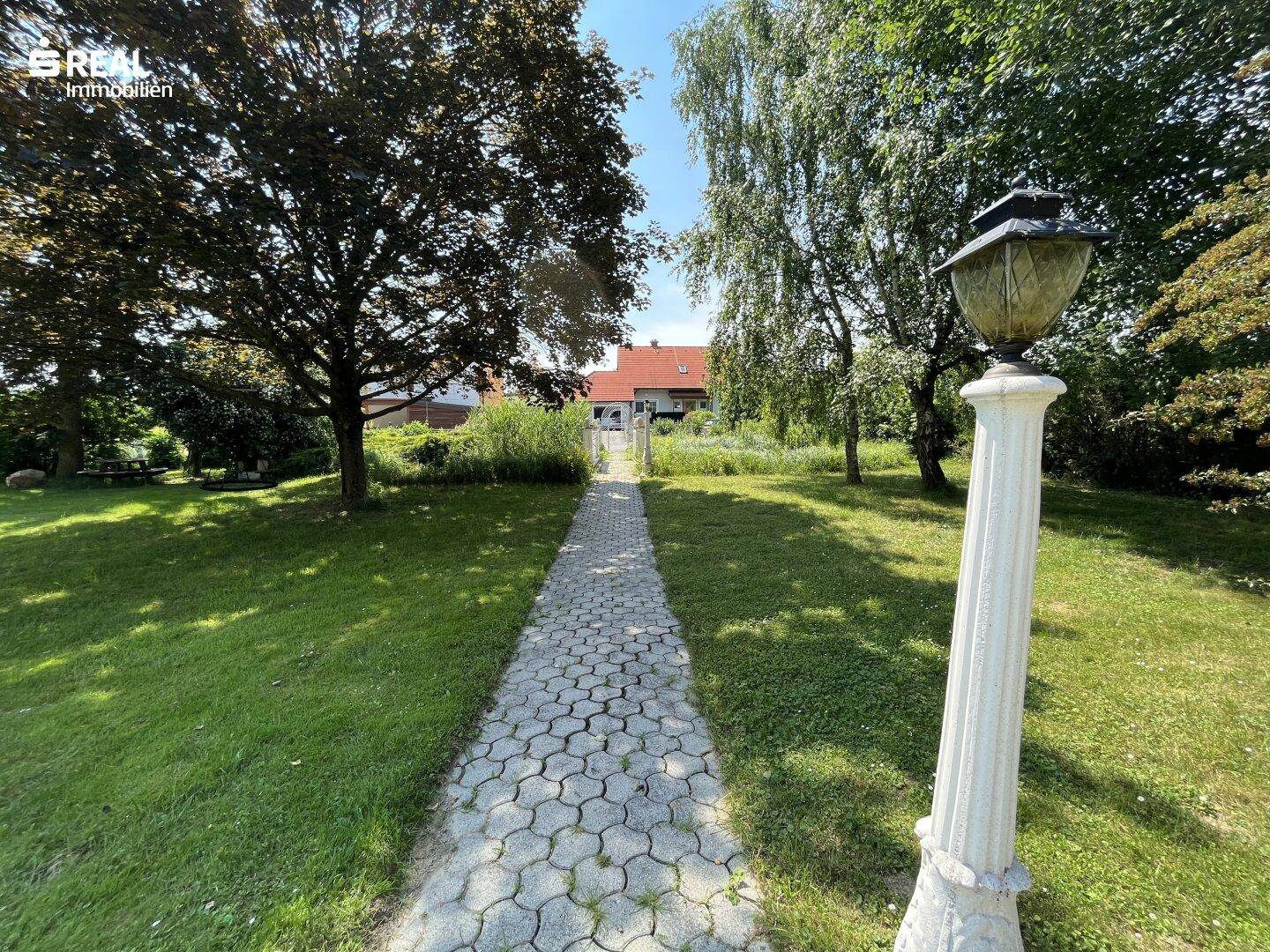 Geschwungener Gartenweg aus Pflastersteinen führt zum Haus, flankiert von Rasen und einem dekorativen Laternenpfahl.