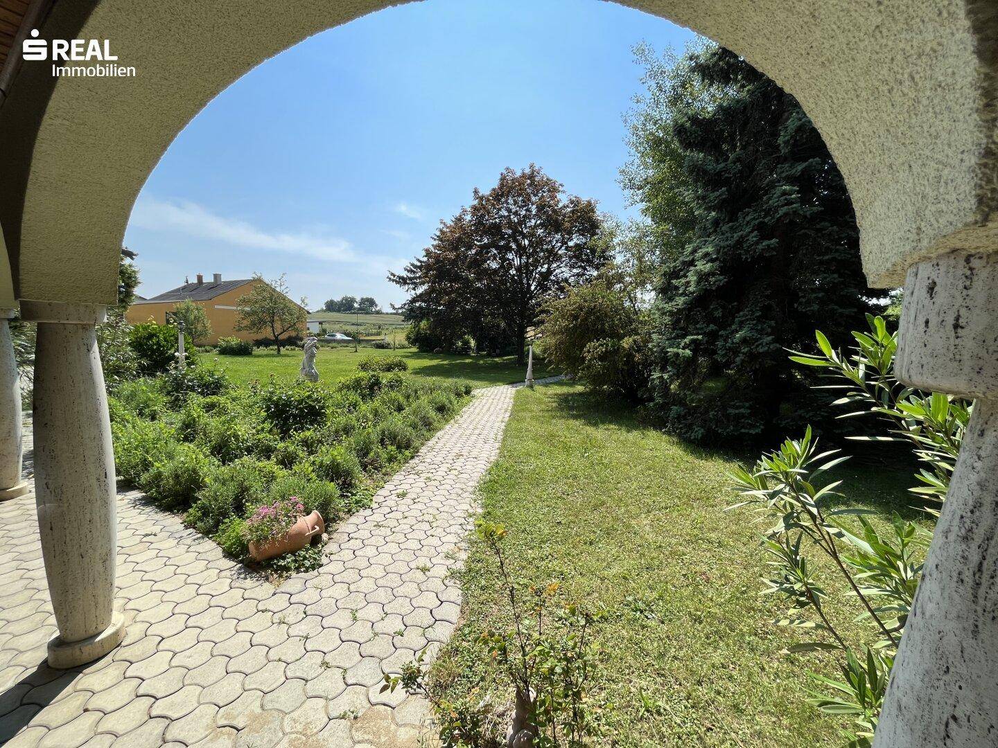 Blick durch einen eleganten Bogen auf den weitläufigen Garten mit gepflastertem Weg und üppiger Vegetation.